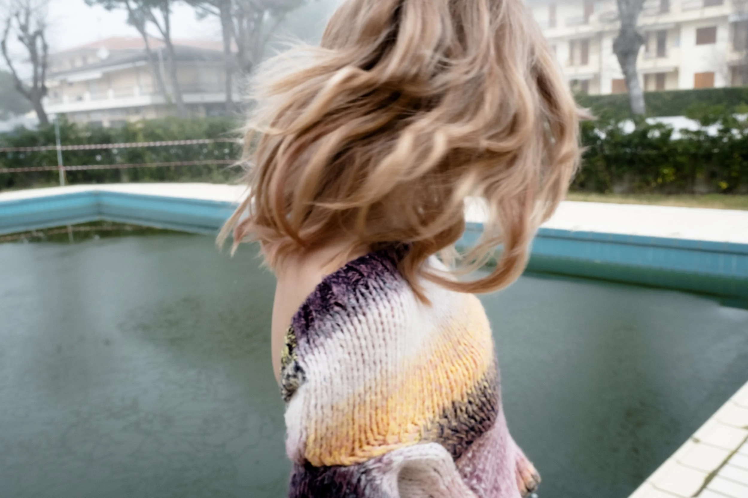 A woman with shoulder-length wavy hair standing near a small pool. She is wearing a colorful, knitted sweater and facing away from the camera. In the background, there are trees and apartment buildings.