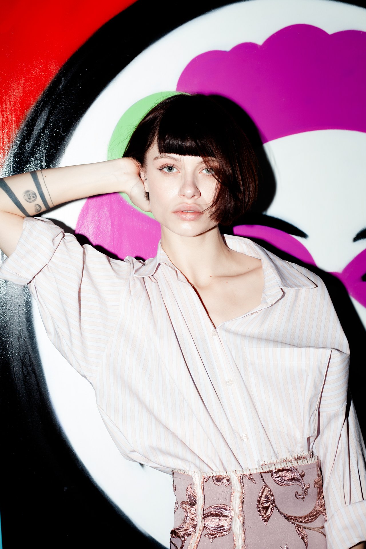 A young woman with short dark hair and pale skin is posing in front of a colorful graffiti background. She is wearing a light pink striped shirt and a skirt with a floral pattern. Her left arm is raised behind her head, revealing tattoos on her forea
