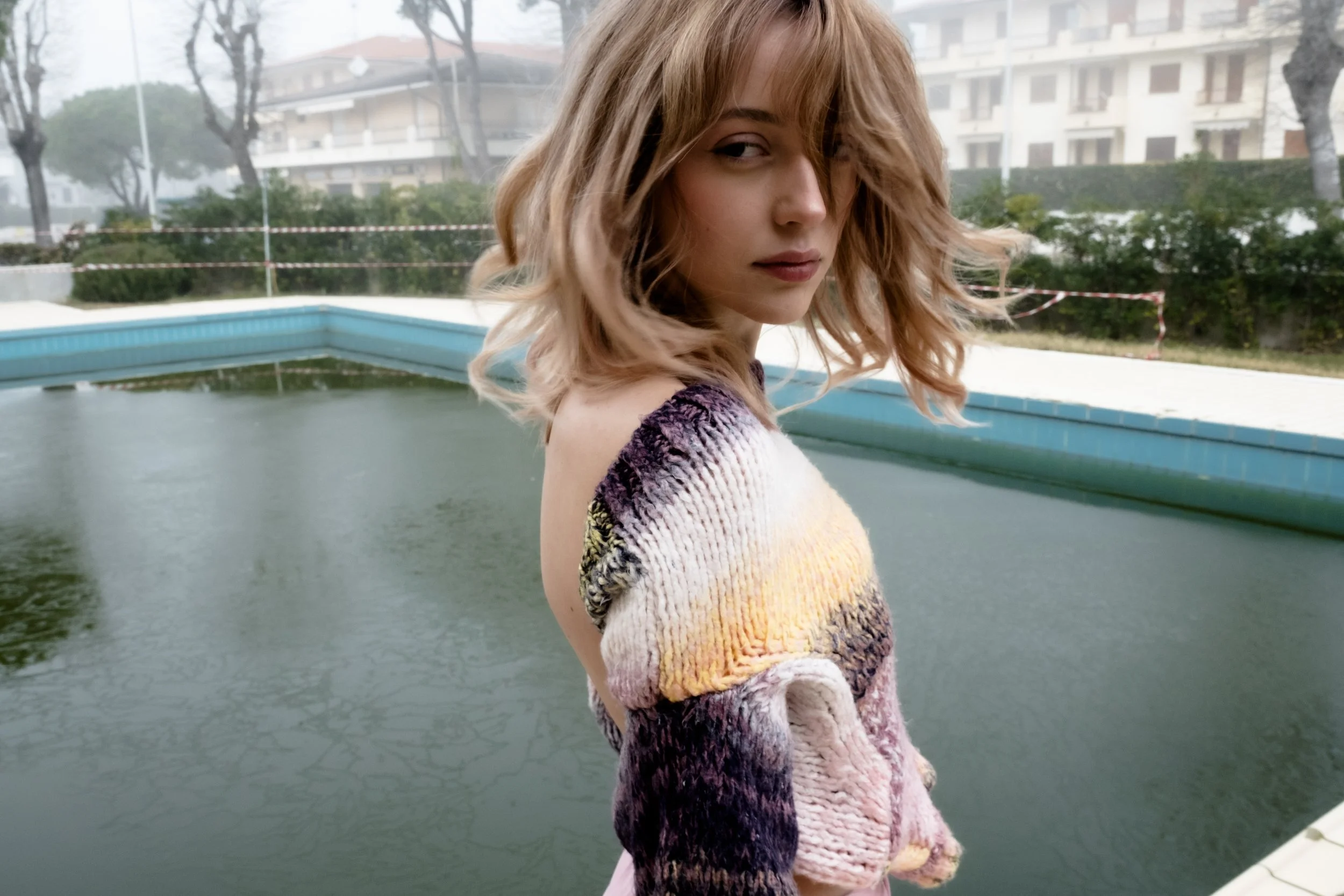 A young woman with shoulder-length wavy blonde hair wearing a multicolored knit sweater standing outdoors near a small pool or pond, with trees and residential buildings in the background.