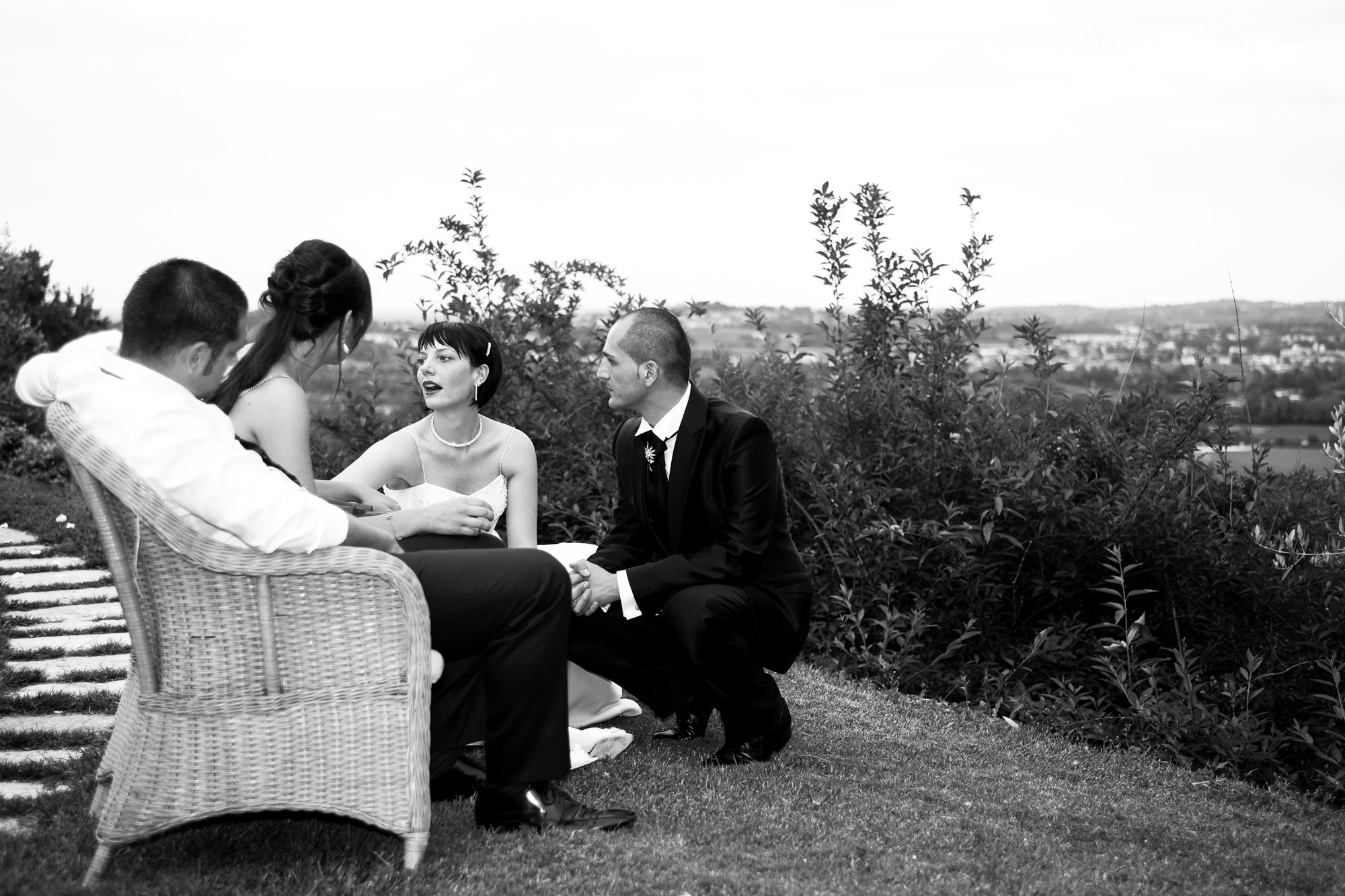 A black and white photo of a wedding ceremony outdoors, with five people sitting and kneeling on the grass, including the bride and groom at the center, with a scenic landscape in the background.