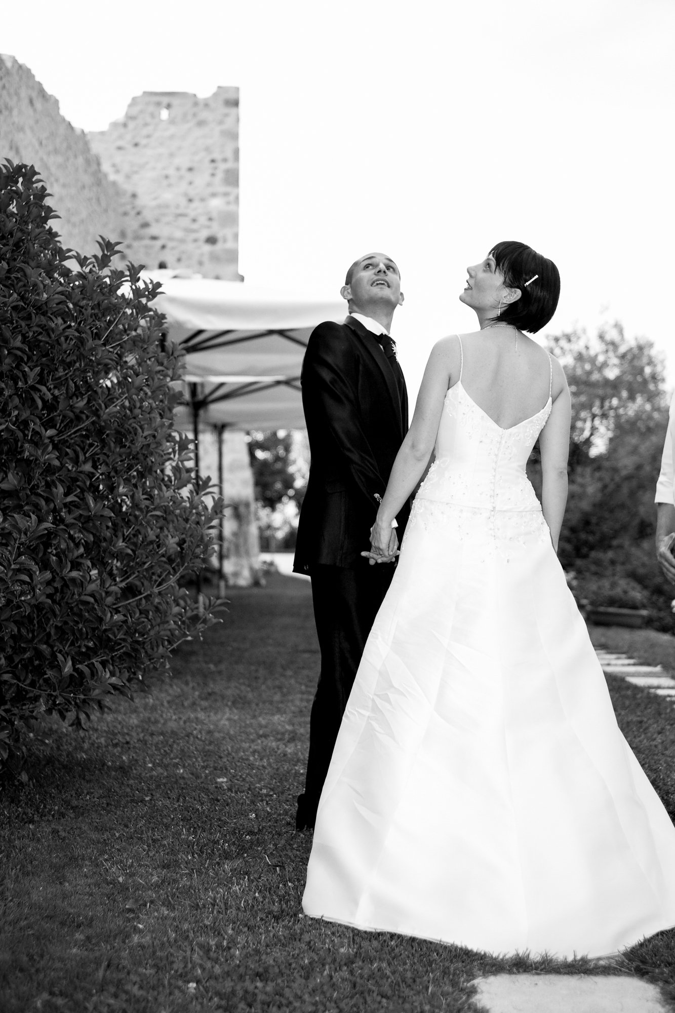 Black and white photo of a couple holding hands outdoors during a wedding, with the woman in a white wedding dress and the man in a dark suit, looking upwards.