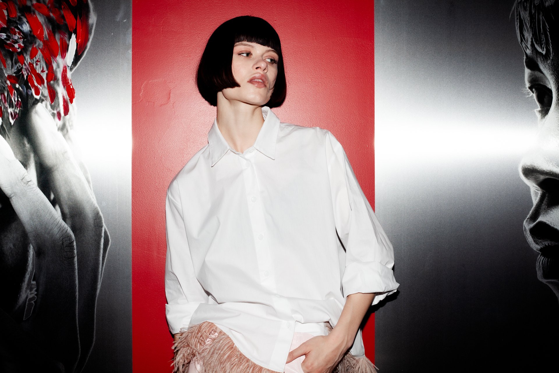 A woman with a short black bob haircut dressed in a white button-up shirt, standing in front of a red background flanked by black and white images of faces.