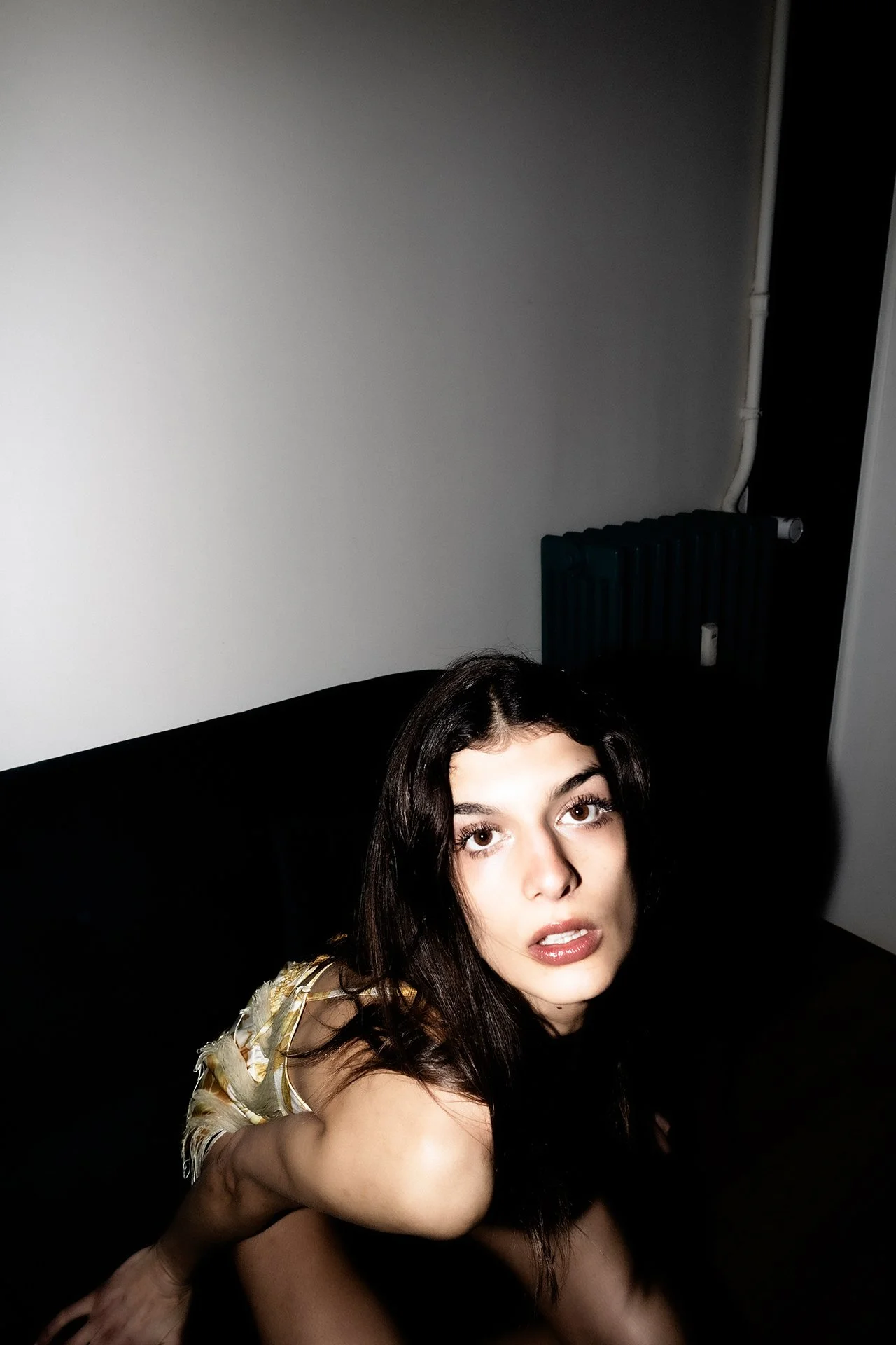 A woman with long dark hair sitting on a black couch in a dimly lit room, looking directly at the camera with a neutral expression.