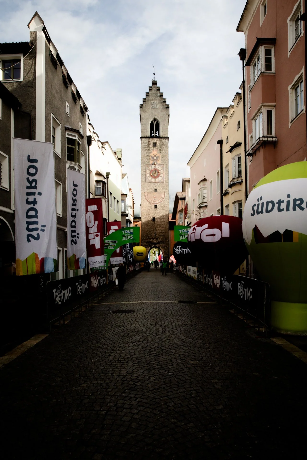 Street view with a historic clock tower at the end, pavement path, and colorful banners and balloons related to cycling events on both sides.
