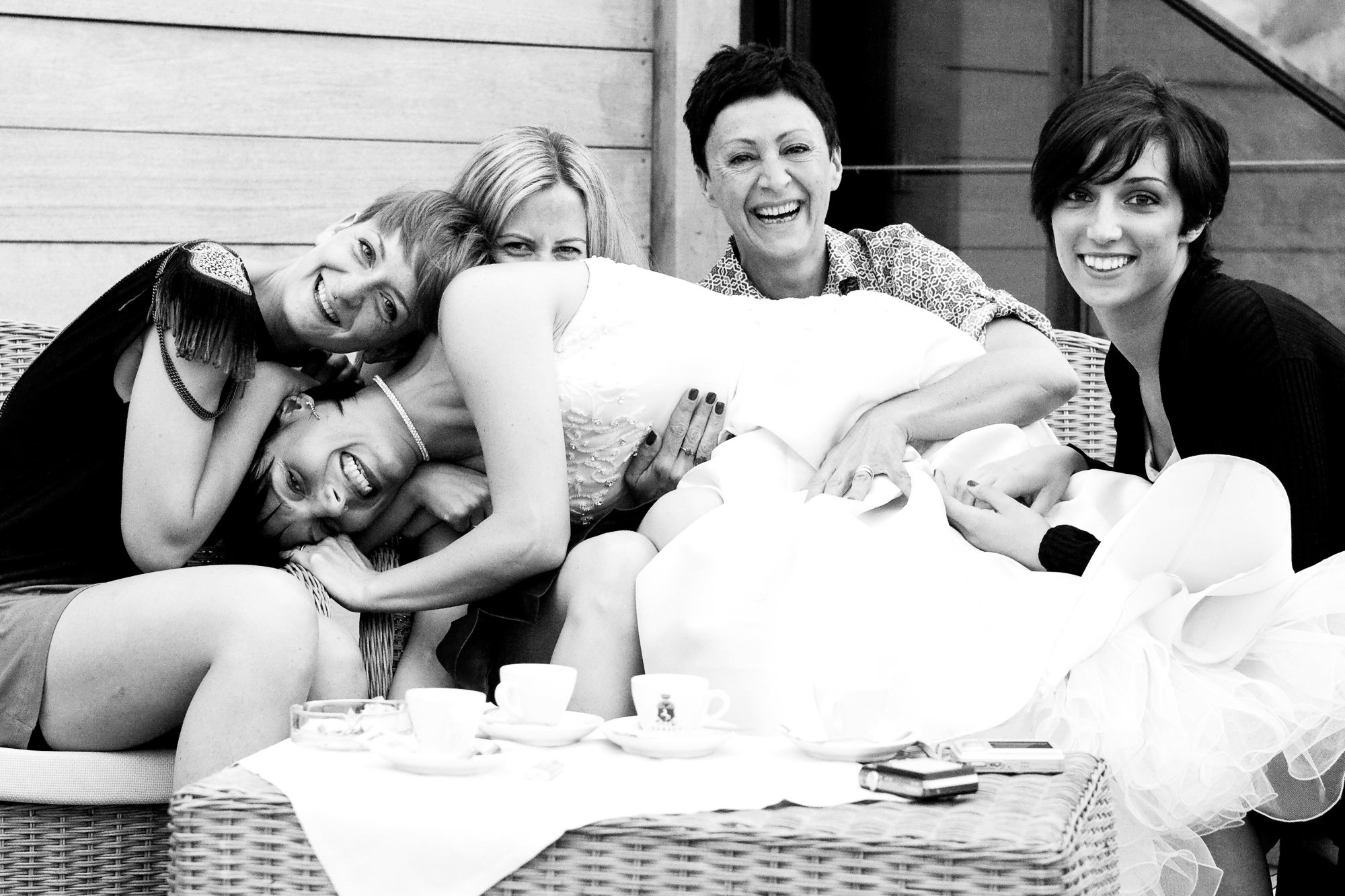 Group of five women, smiling and lying close together on a couch, with one woman in a wedding dress, celebrating at a social gathering.