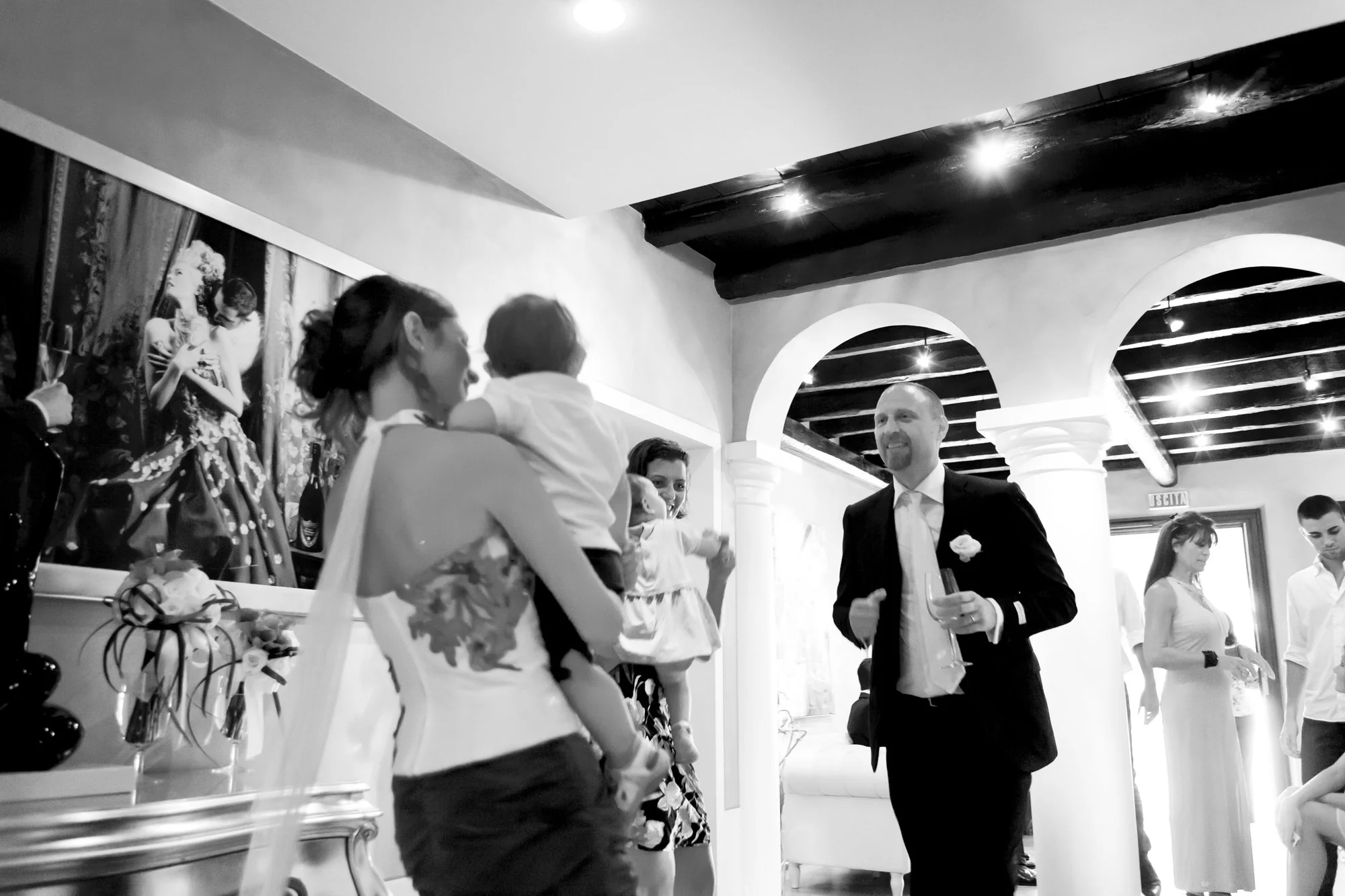 Black and white photo of a wedding celebration in a decorated room with arches. A man in a suit is holding a drink and talking to a woman holding a child, both smiling. Other guests are in the background.