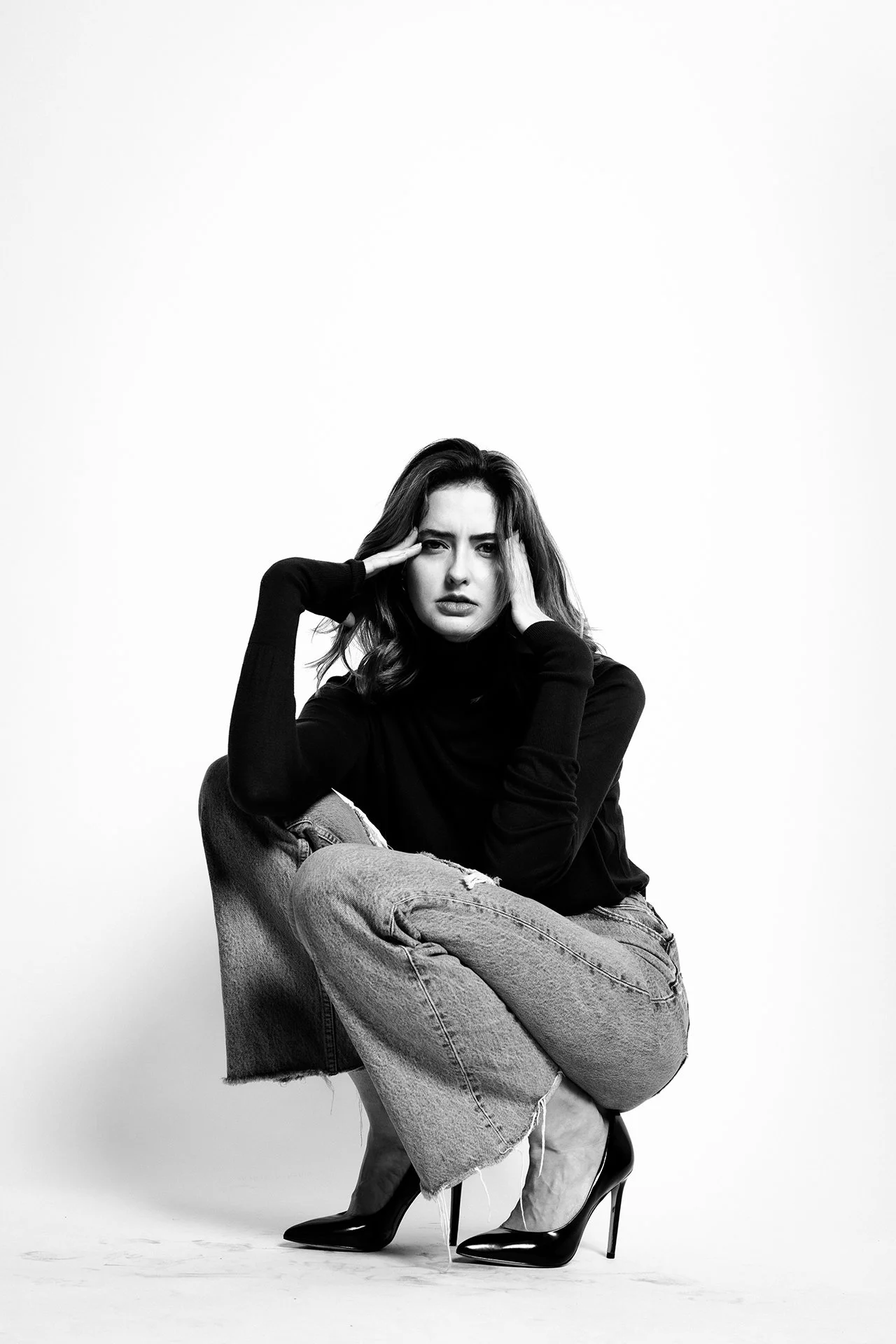 Black and white photo of a woman with dark wavy hair in a black turtleneck and ripped jeans, wearing high heels, sitting on the ground with one knee up, holding her head with her hands, and looking at the camera.