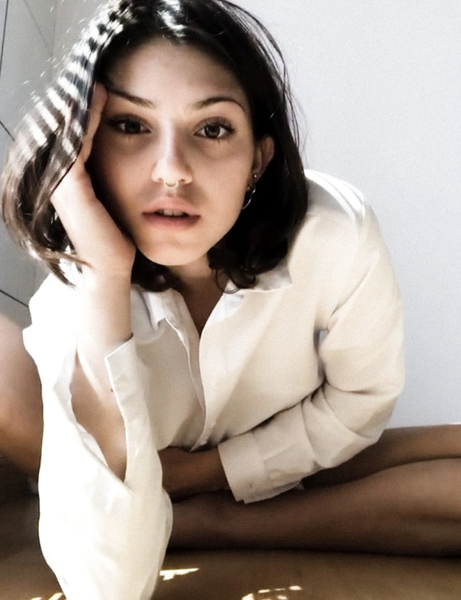 A young woman with dark hair and earrings, wearing a white shirt, sitting on the floor with her legs crossed, looking into the camera with a surprised expression, hand resting on her cheek.