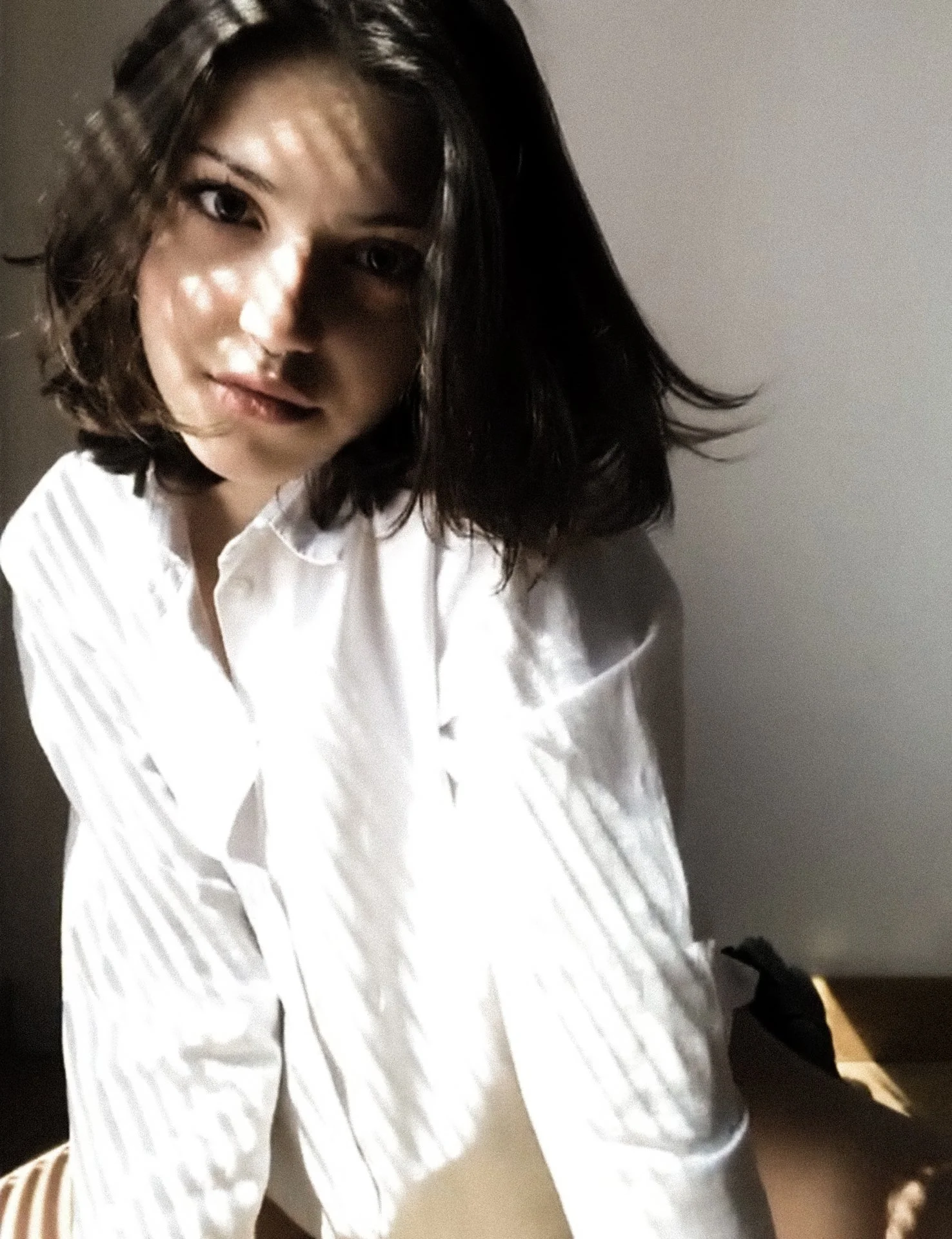 A woman with shoulder-length dark hair and a white shirt, sitting against a plain wall, with sunlight casting shadow patterns across her face.