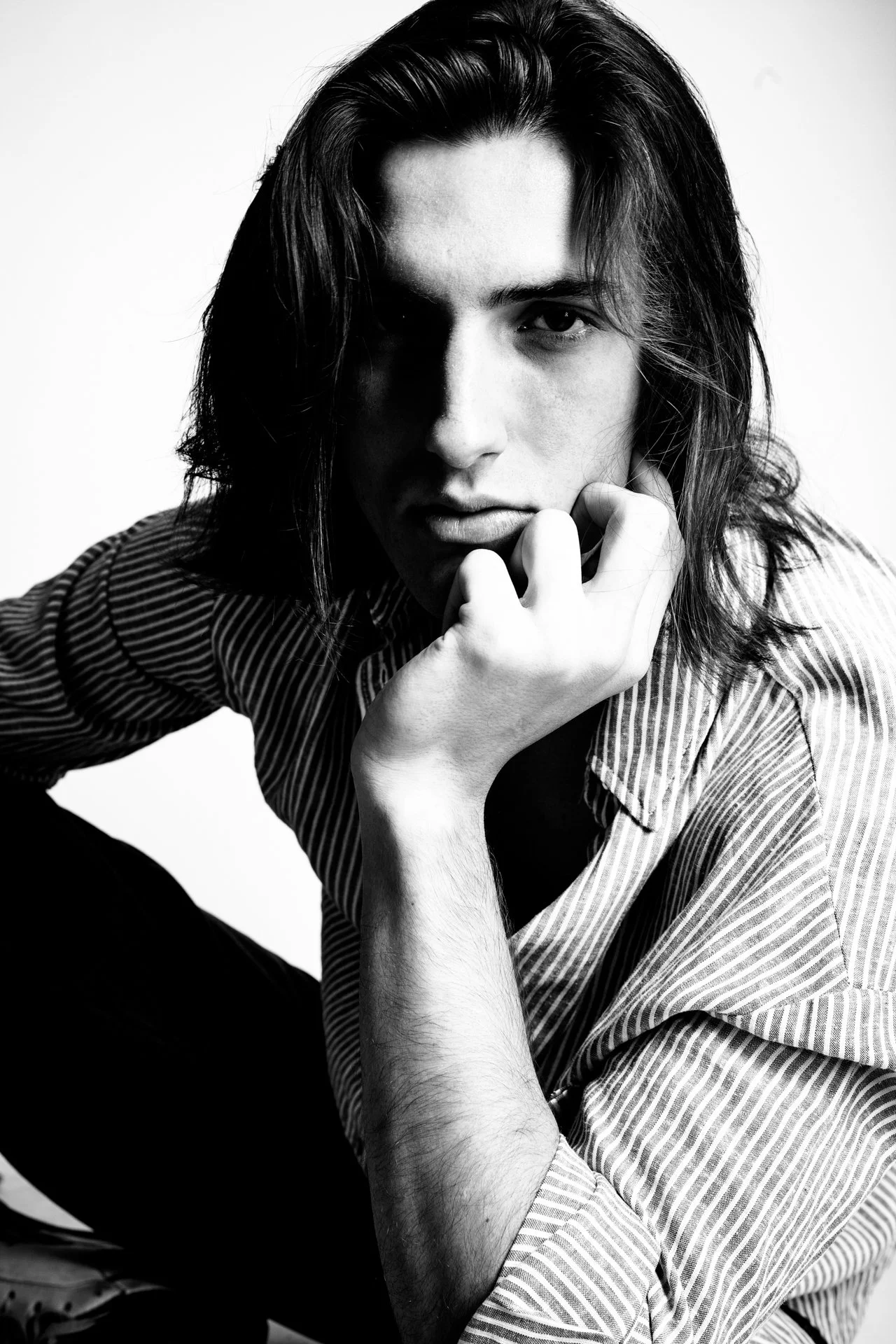 Black and white portrait of a young man with long hair, wearing a striped shirt, looking intently at the camera with his hand resting on his chin.