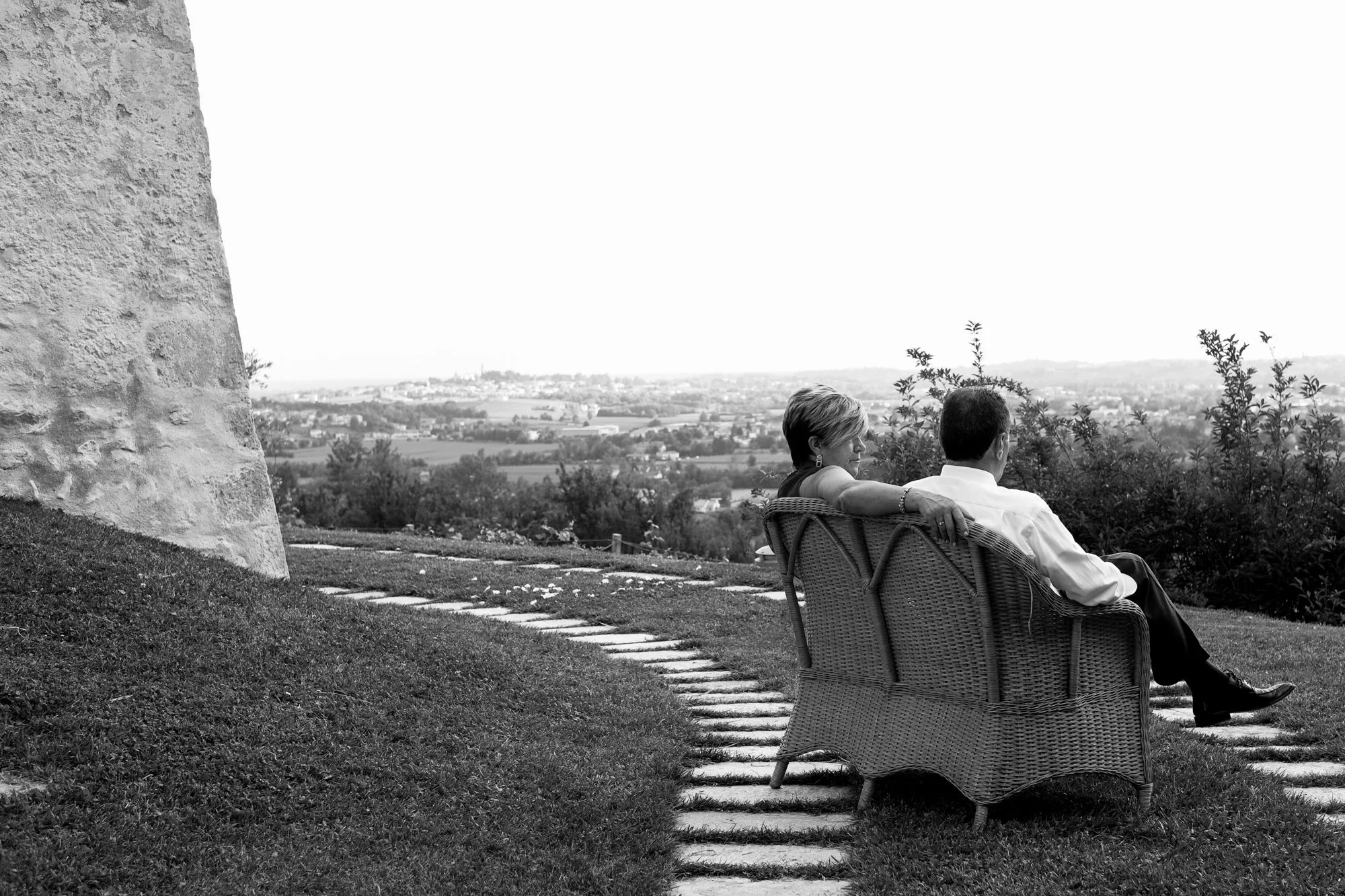 A man and woman sitting on a wicker bench on a grassy hill, overlooking a landscape of fields and distant buildings.