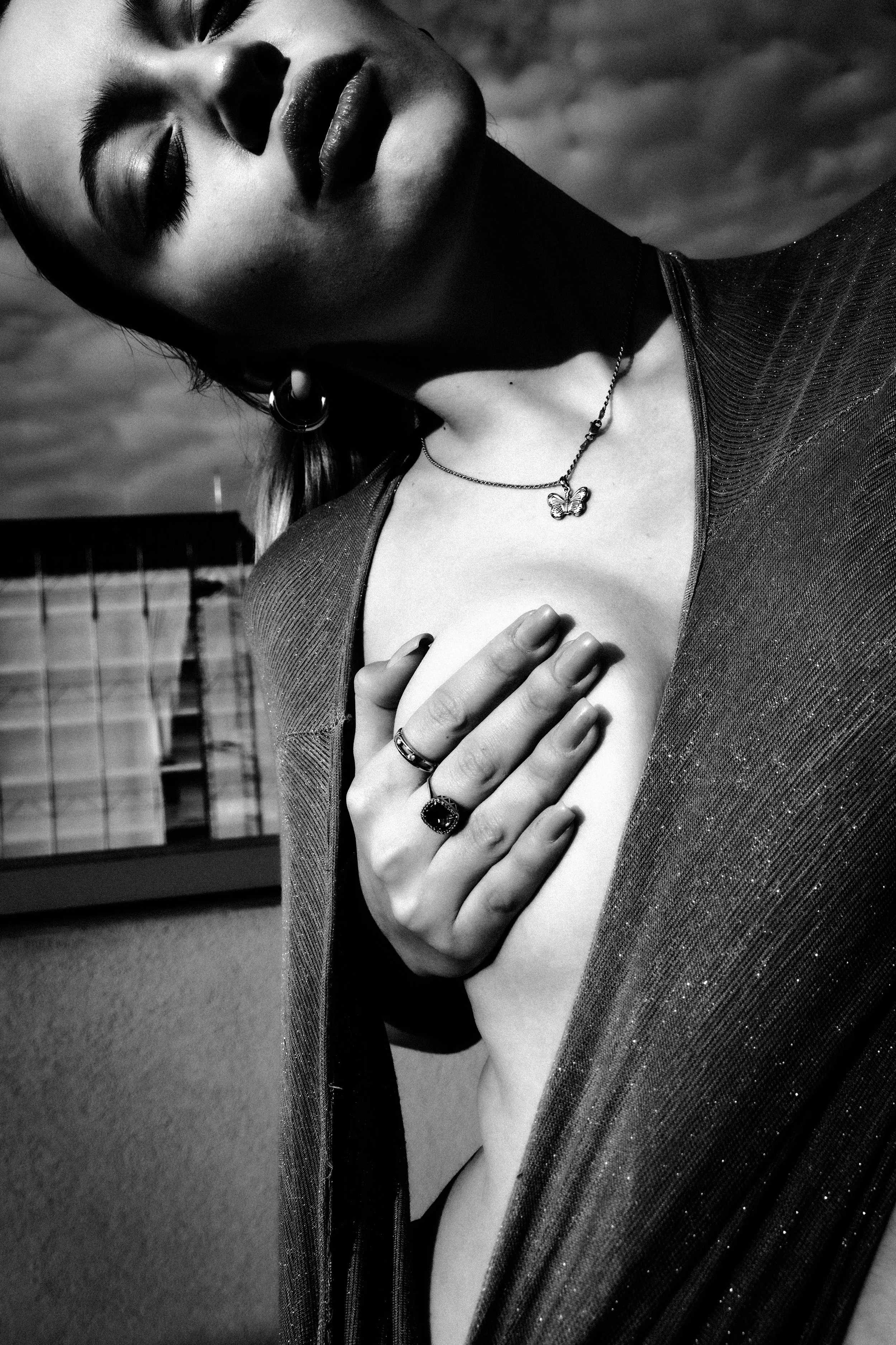 A black-and-white photo of a woman with short hair, closed eyes, and makeup, holding her hand over her chest. She wears a necklace with a butterfly pendant, hoop earrings, and rings, and is dressed in a shimmering garment. The background features a b