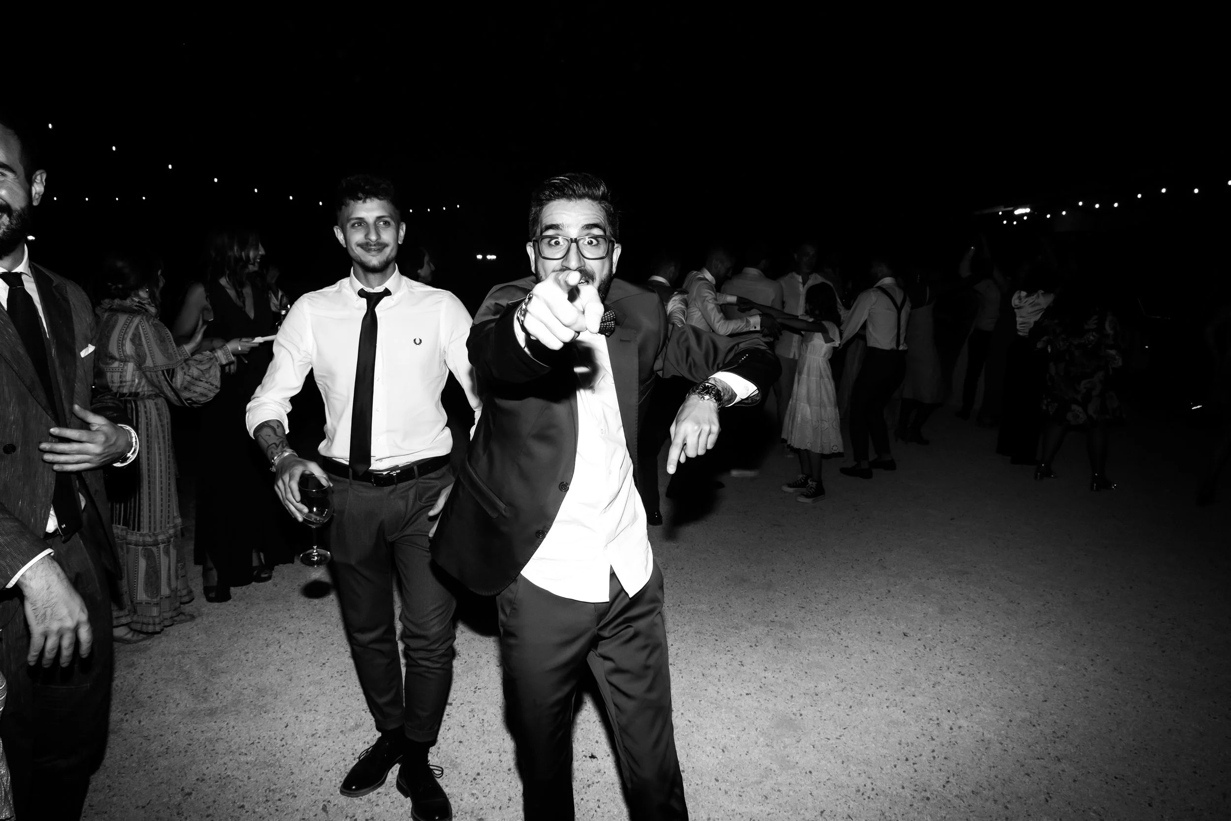 A black and white photo of a man in a tuxedo and glasses pointing at the camera, holding a drink in his other hand, at a lively social event with other well-dressed people dancing and mingling in the background.