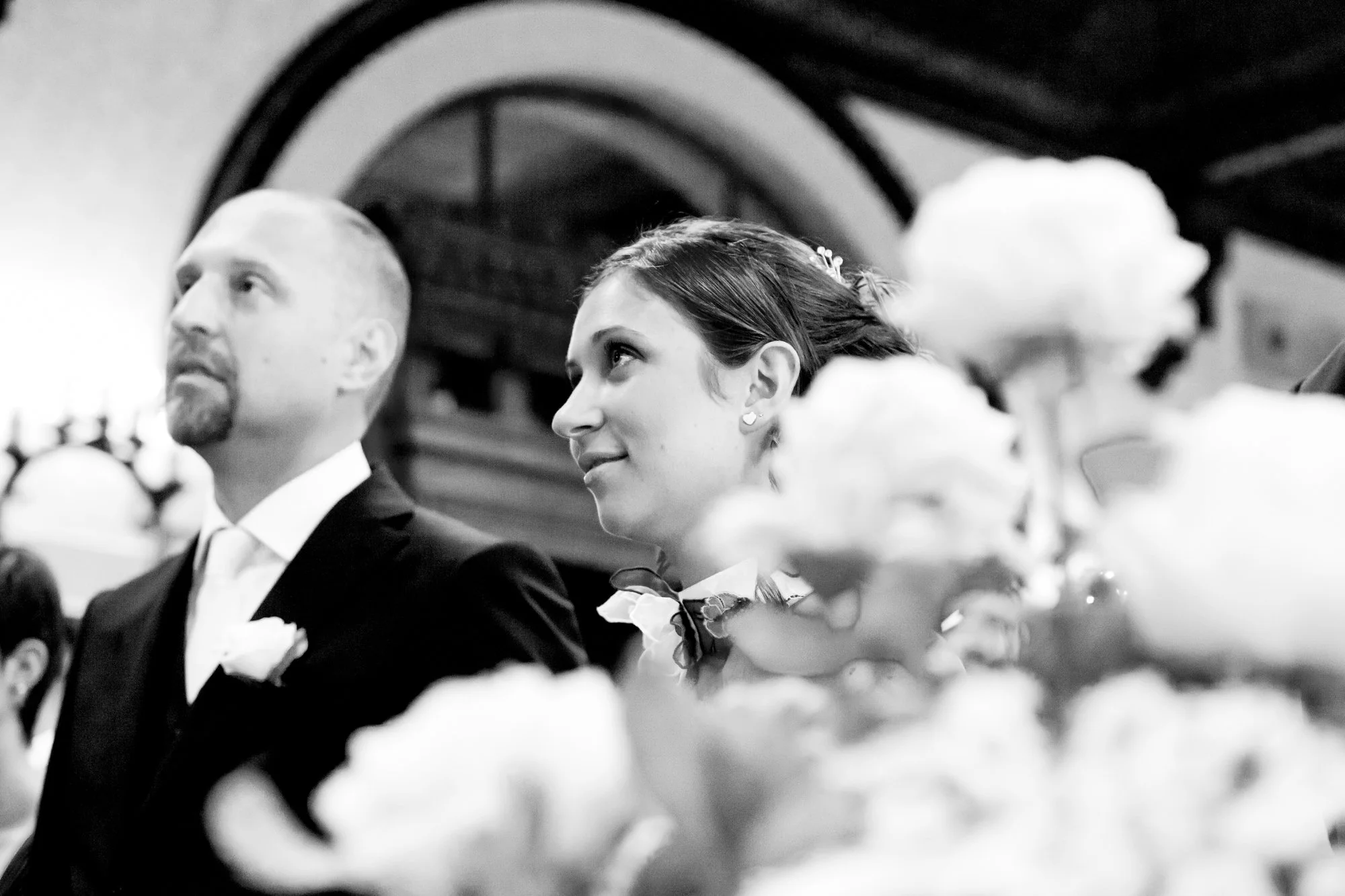 A bride and groom sitting during their wedding ceremony in a church, with flowers in the foreground and an arched architectural detail in the background.