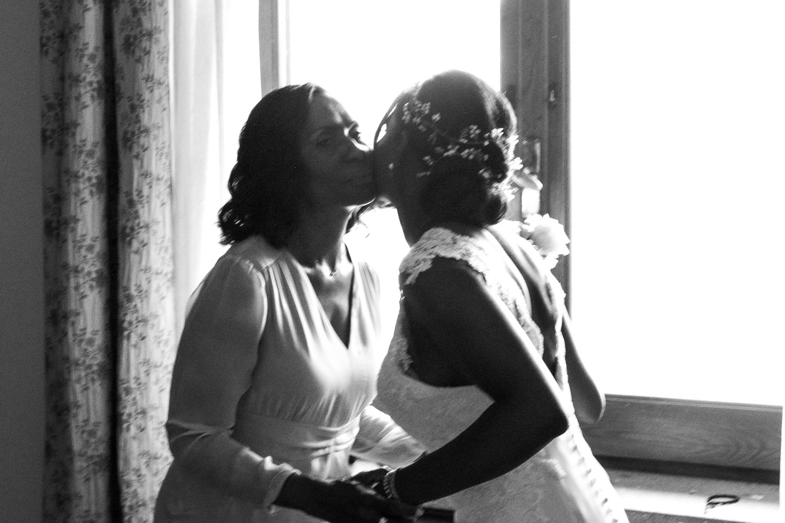 A woman in a wedding dress and another woman, possibly her mother, sharing a kiss in front of a window.