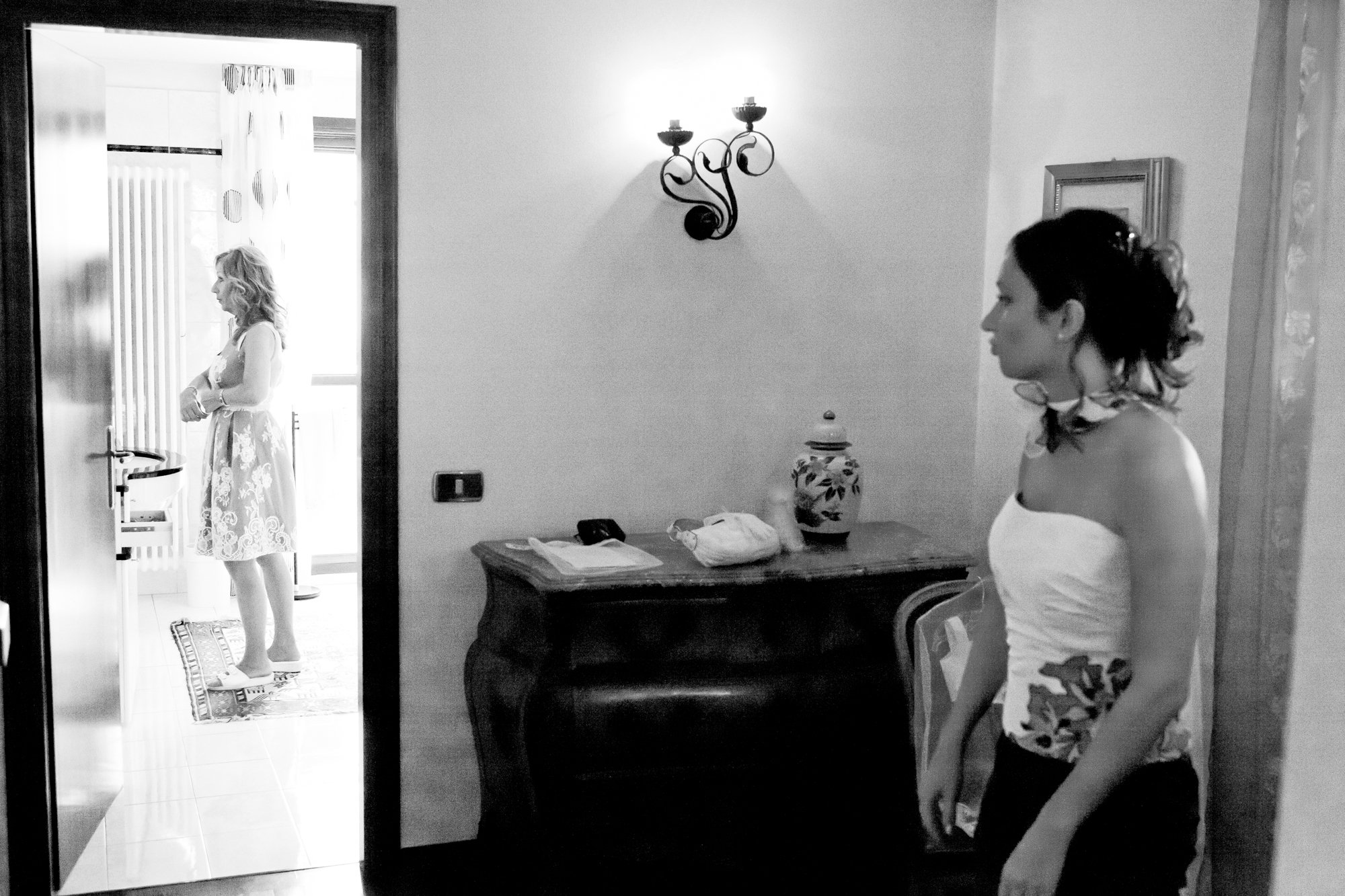 Two women in a room; one standing in a doorway, looking out, wearing a floral dress, and the other standing near a wall, wearing a strapless top and floral scarf, with their profile visible.