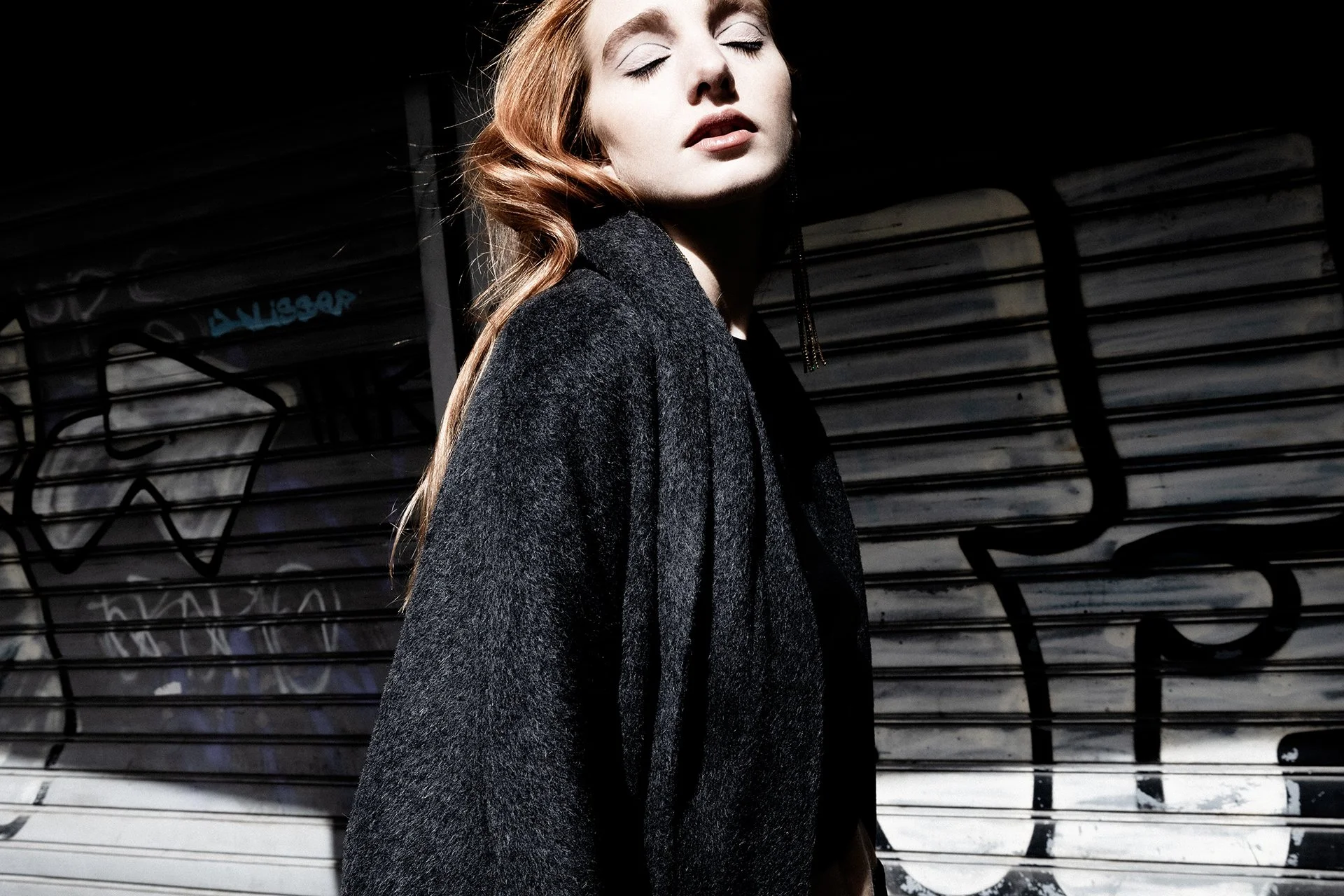 A woman with closed eyes and red hair styled in loose waves, wearing a dark coat, standing against a graffiti-covered metal shutter.