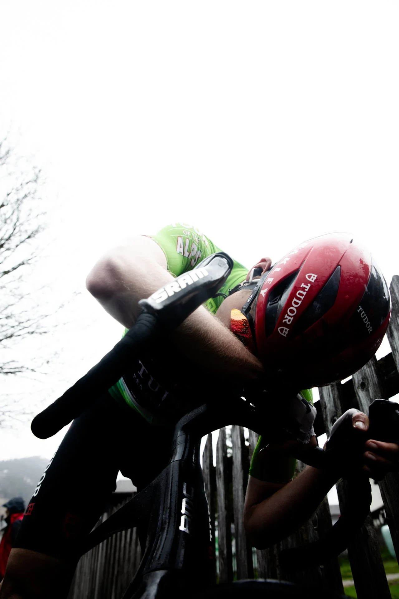 A cyclist wearing a red helmet and green jersey leaning on a black bicycle, with a wooden fence in the background and overcast sky.