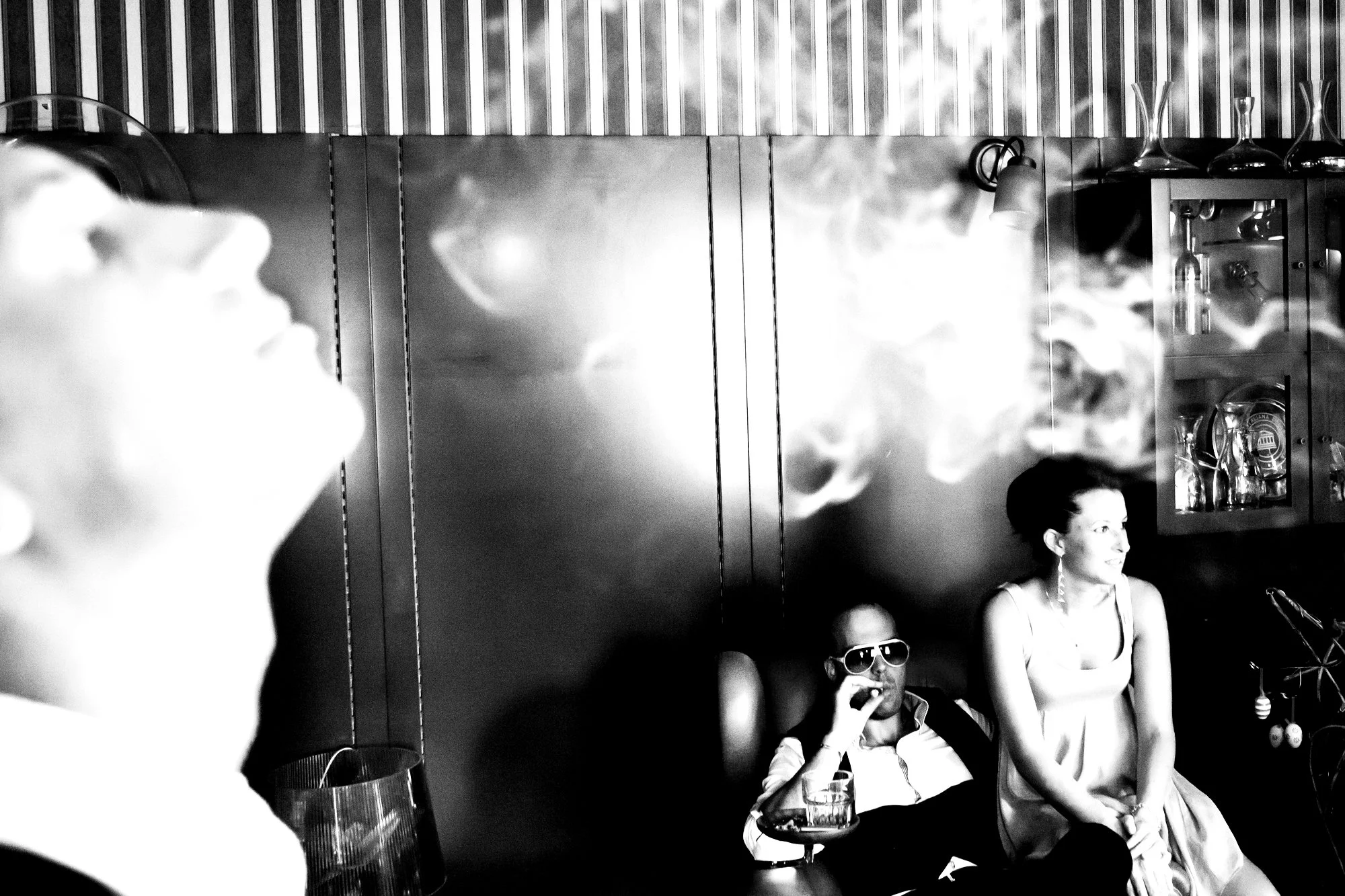 Black and white photo of three people sitting indoors. On the left, part of a person's face with their eyes closed and head tilted back, out of focus. In the middle, a man wearing sunglasses, smoking, and holding a drink. On the right, a woman smilin