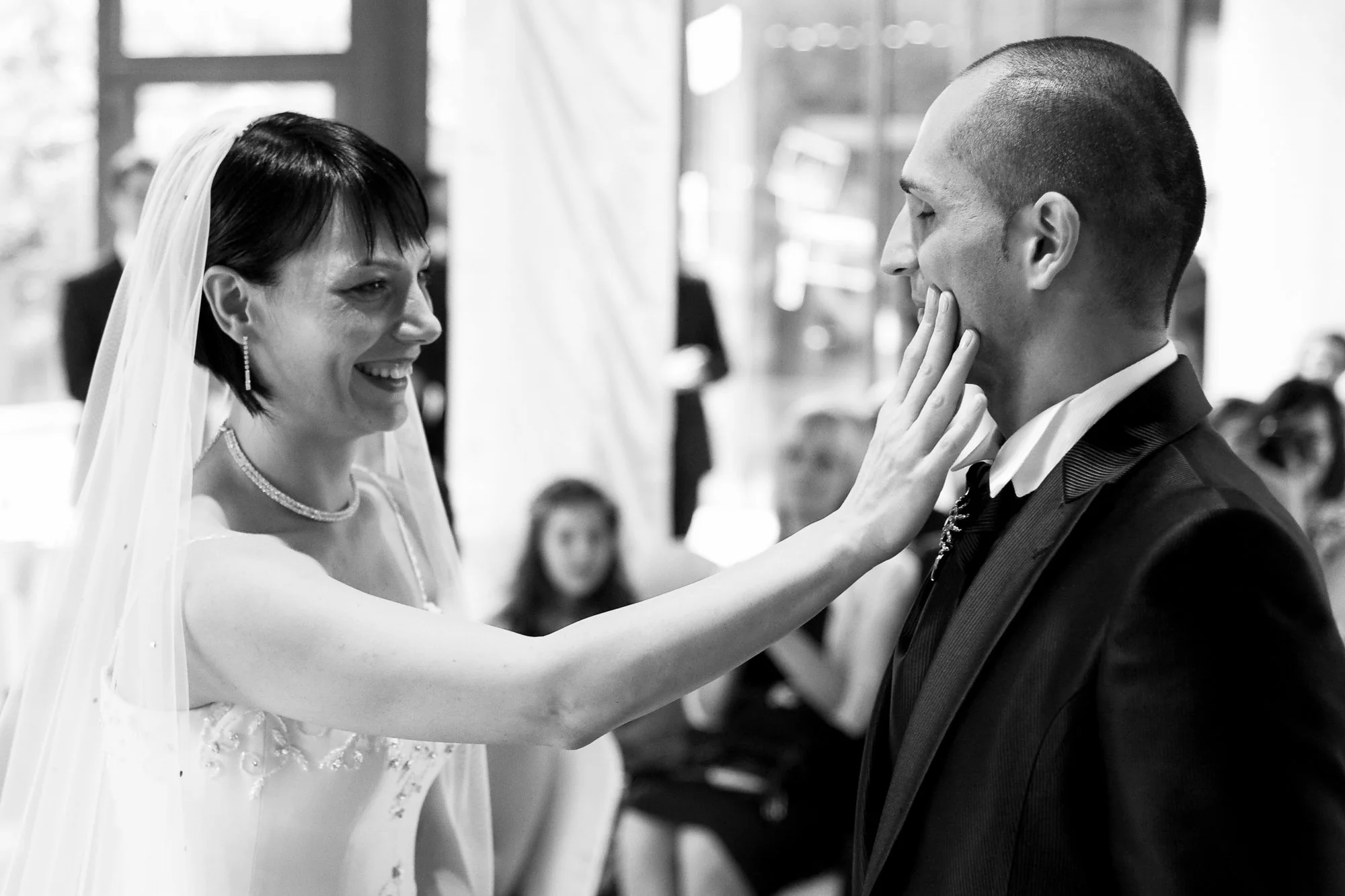 A bride smiling as she touches her groom's face during their wedding ceremony, both dressed in formal attire and surrounded by guests.