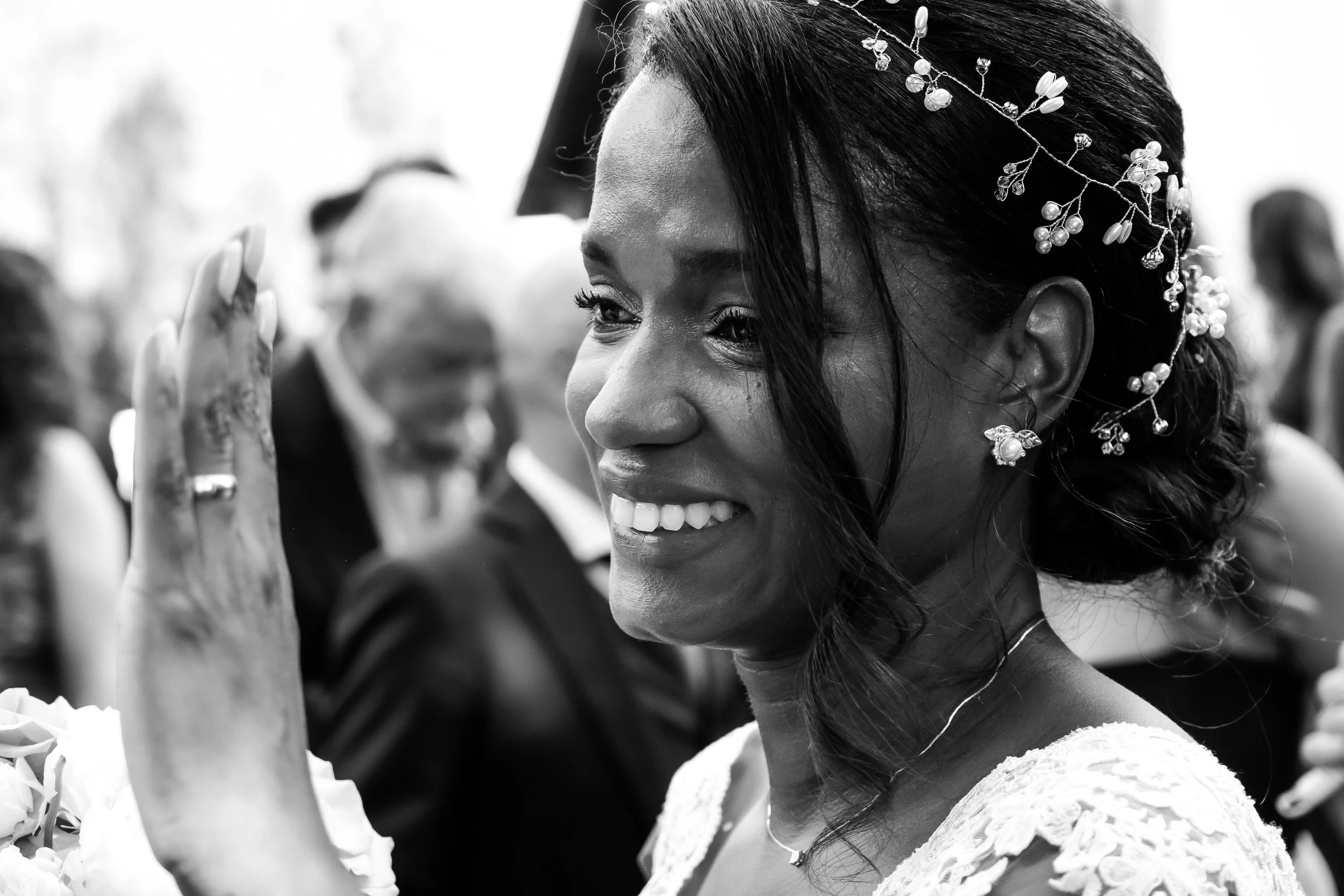 Black-and-white photo of a smiling woman with dark hair styled with a decorative hairpiece, wearing earrings and a lace dress, waving her hand during a celebration, with blurred people in the background.