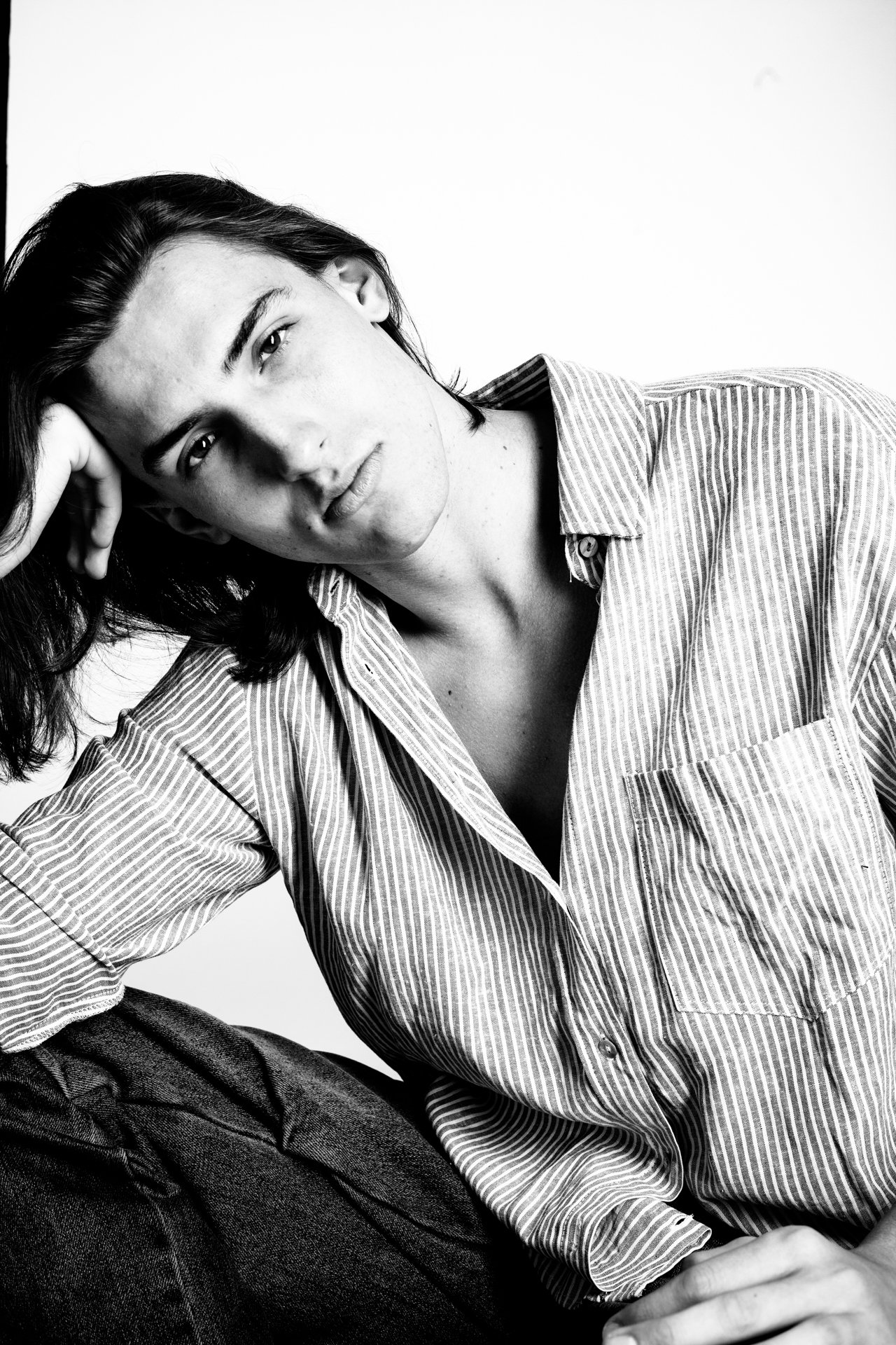 Black and white portrait of a young woman with dark hair, wearing a striped shirt, sitting with her hand resting on her head, against a plain background