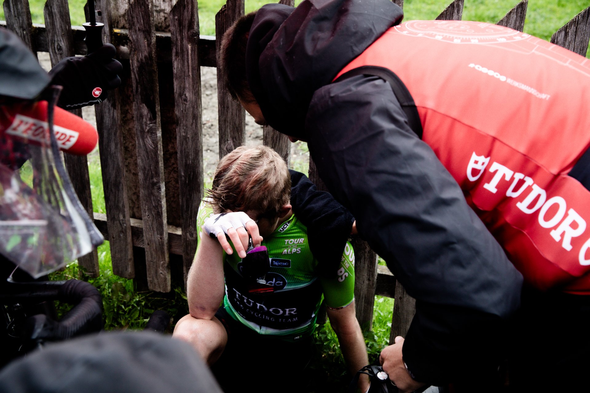 A cyclist sits on the ground, being attended to by a team member wearing a red jacket with Tudor branding, after a crash or fall during a cycling event. The cyclist is wearing a green racing jersey, and the scene takes place next to a wooden fence.