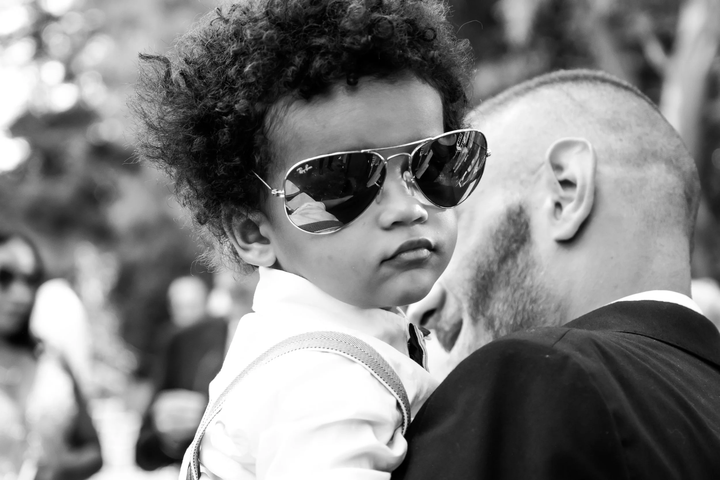 A young child with curly hair wearing reflective sunglasses is being carried by an adult with a shaved head and beard, in an outdoor setting with blurred figures in the background.