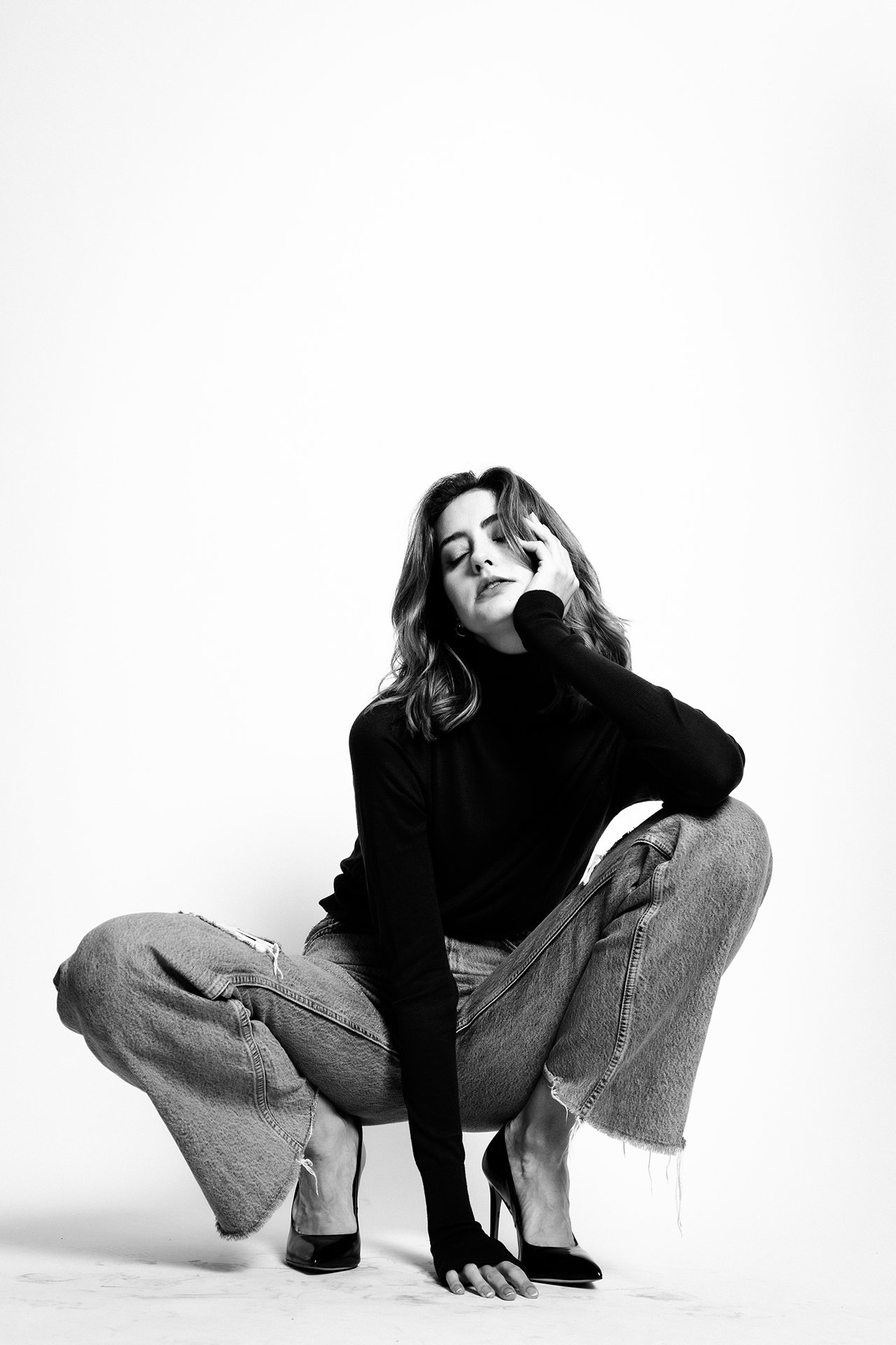 Black and white photo of a woman crouching on the floor, wearing high heels, ripped jeans, and a black top, with one hand touching her face and the other on the ground.