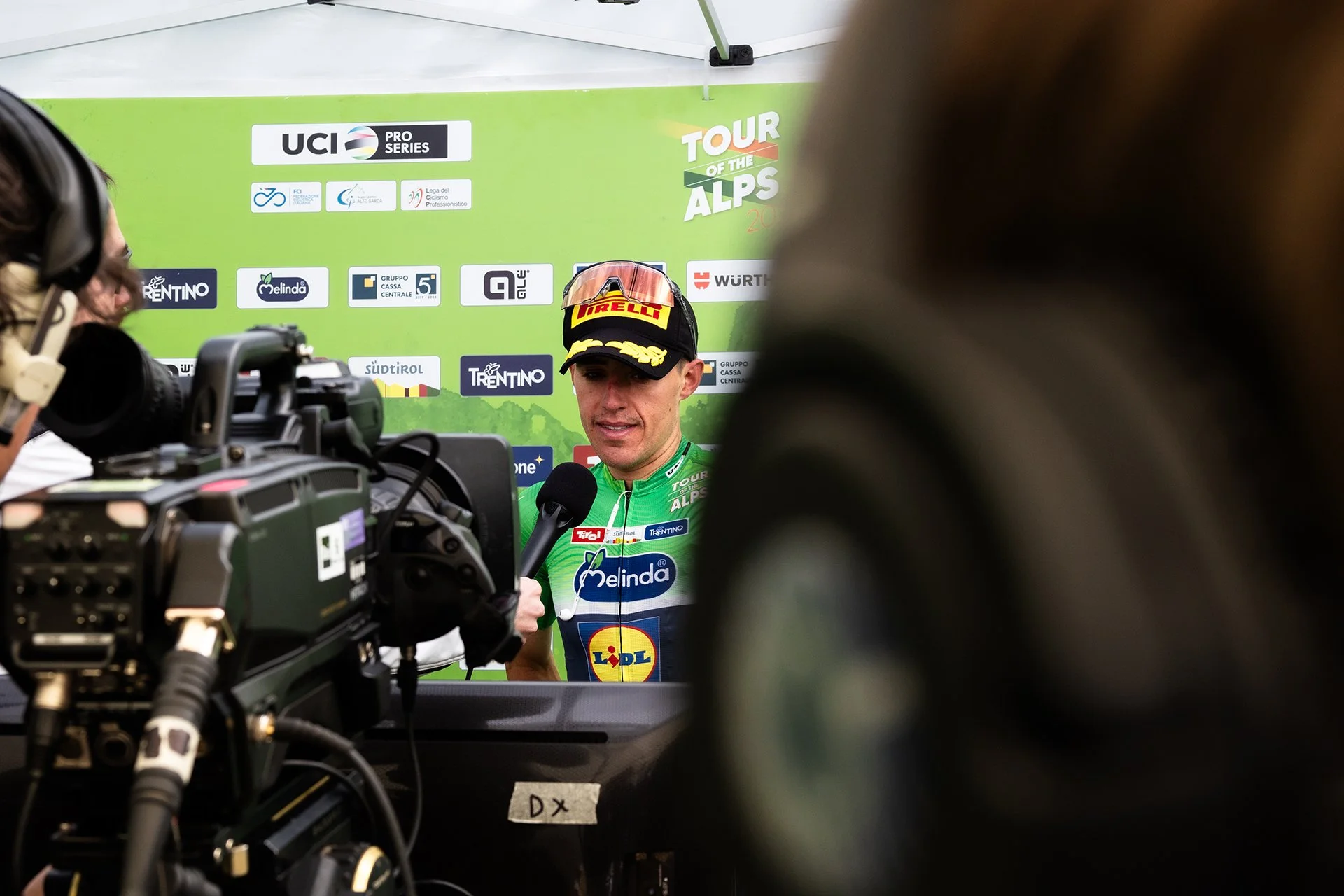 Speaer being interviewed at the Tour of the Alps cycling race, with a green backdrop displaying various sponsor logos.