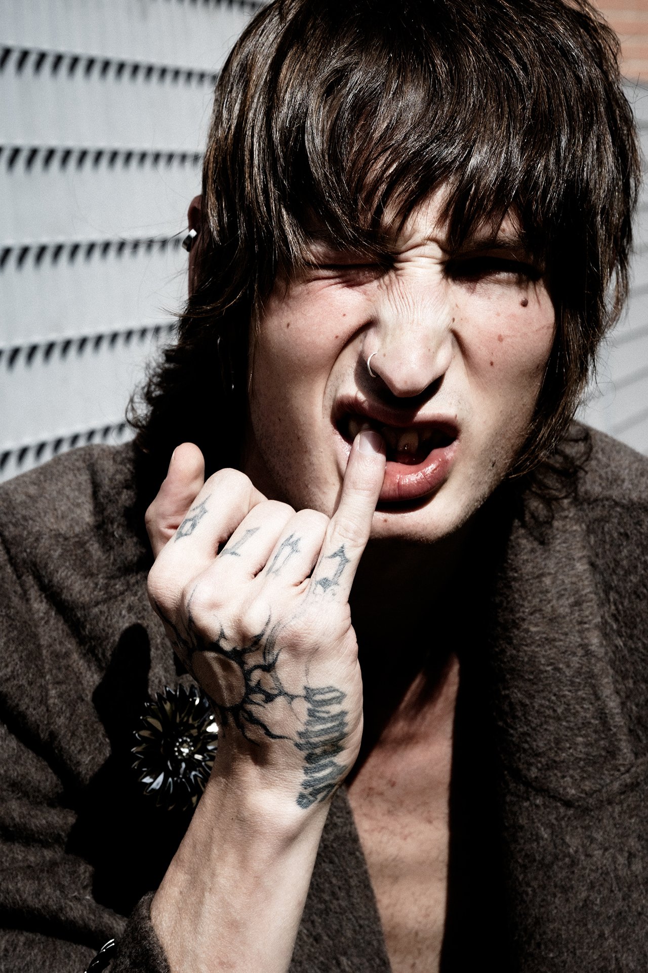 Person with tattoos on hand and face, making a grimace, touching their lip with one finger, in front of a vented metal wall.