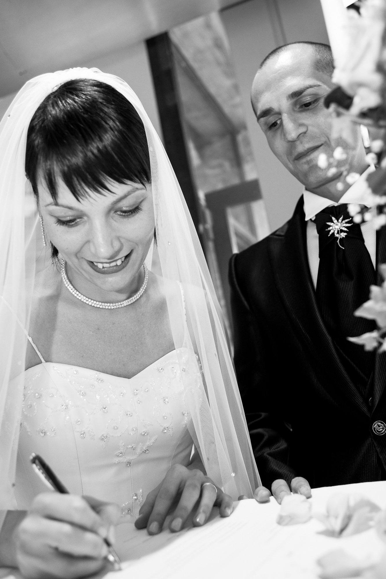 A bride with short dark hair, wearing a veil, pearl necklace, and wedding dress, is signing a document while a groom in a suit looks on at her side.