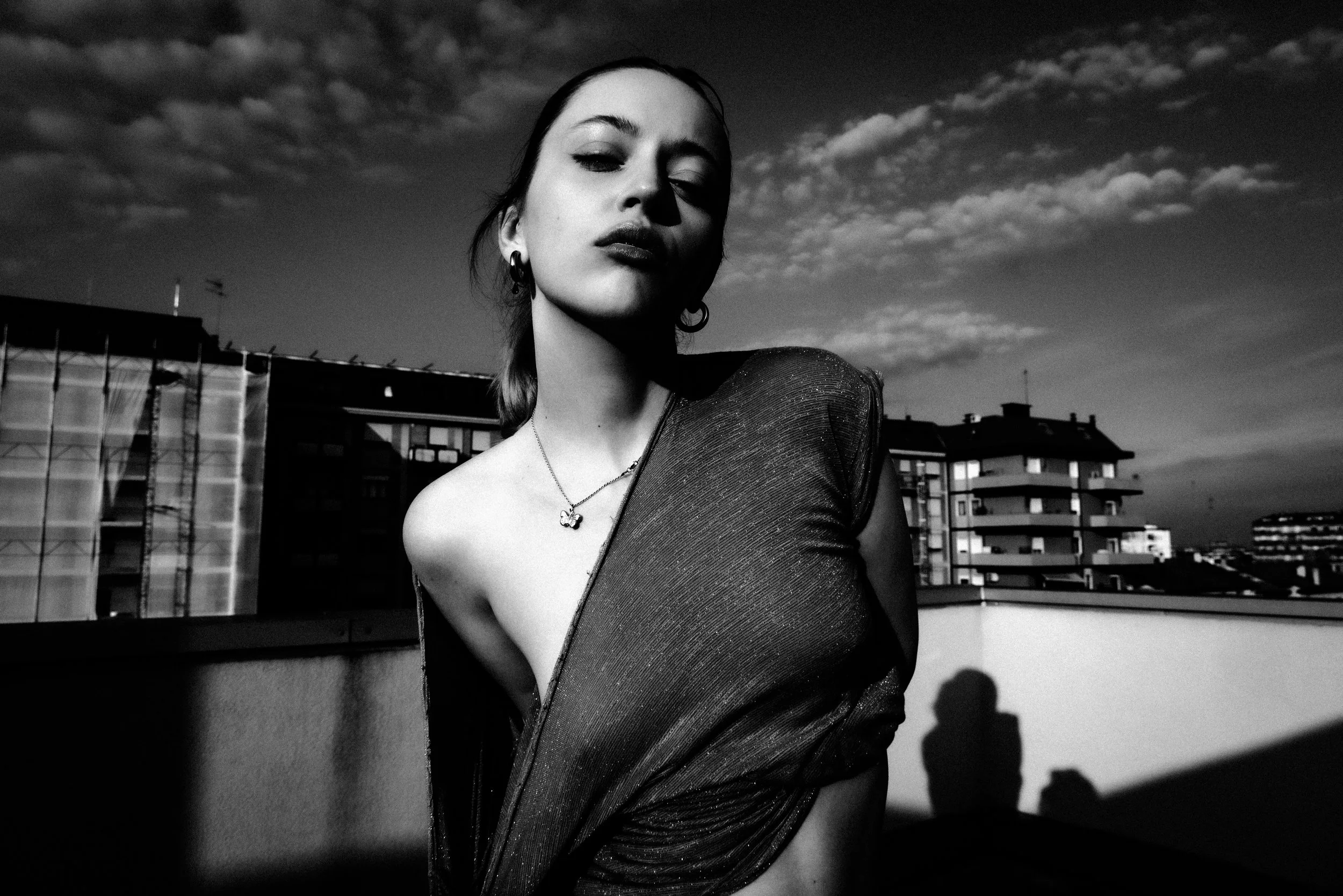 Black and white photo of a woman on a rooftop, wearing a shimmery dress with an asymmetrical neckline, gazing at the camera with partially closed eyes, against a sky with scattered clouds and city buildings in the background.