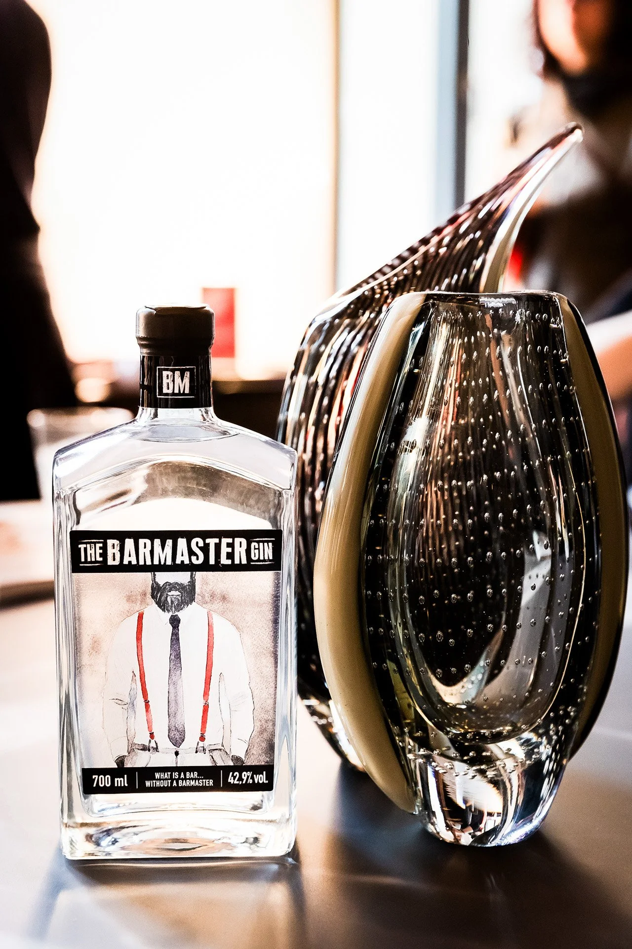 A bottle of The Barmaster Gin on a table, with a unique glass and reflective objects behind it, in a bright indoor setting.