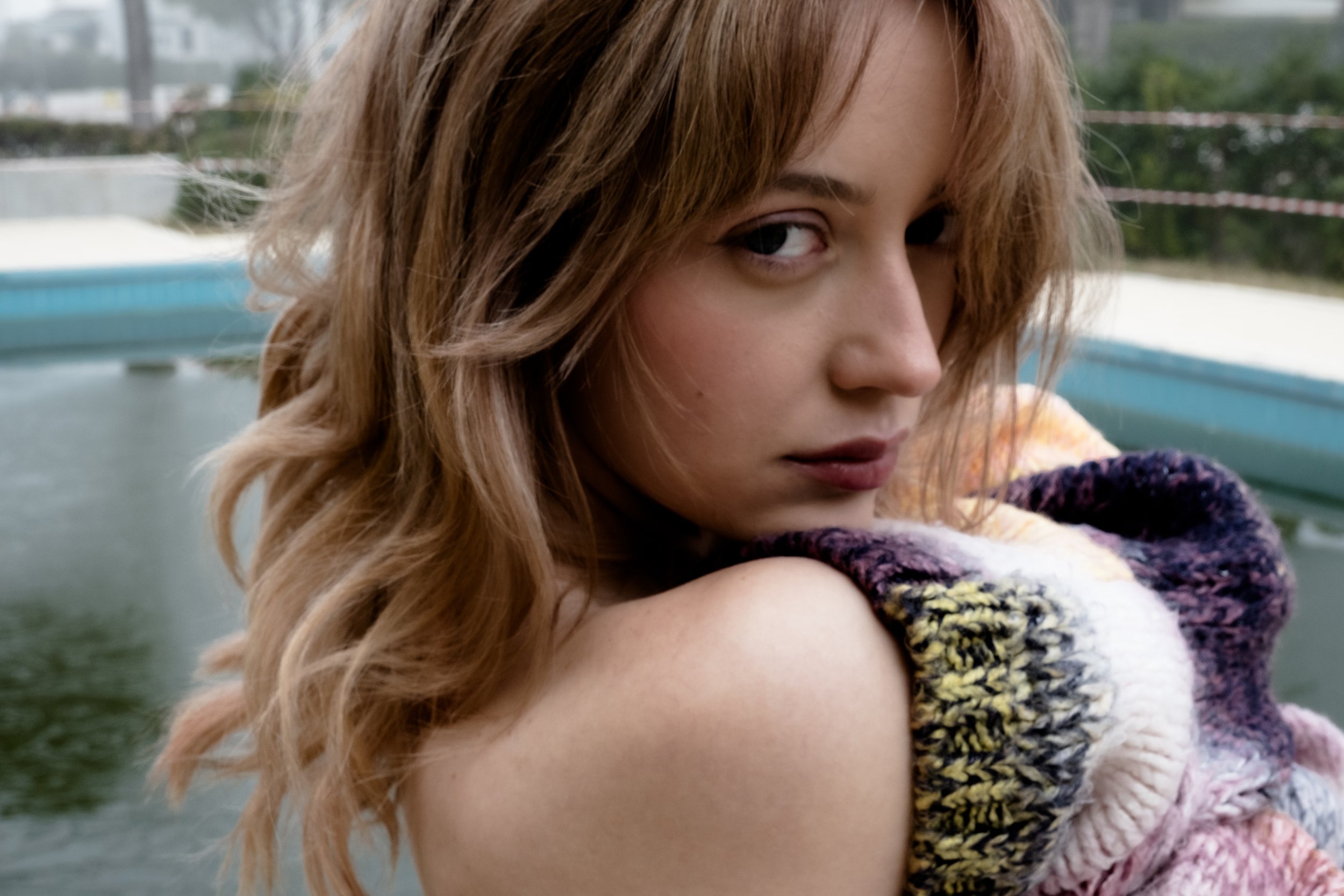 A woman with light brown, wavy hair looking over her shoulder, wearing a colorful knitted sweater, in an outdoor setting near water.