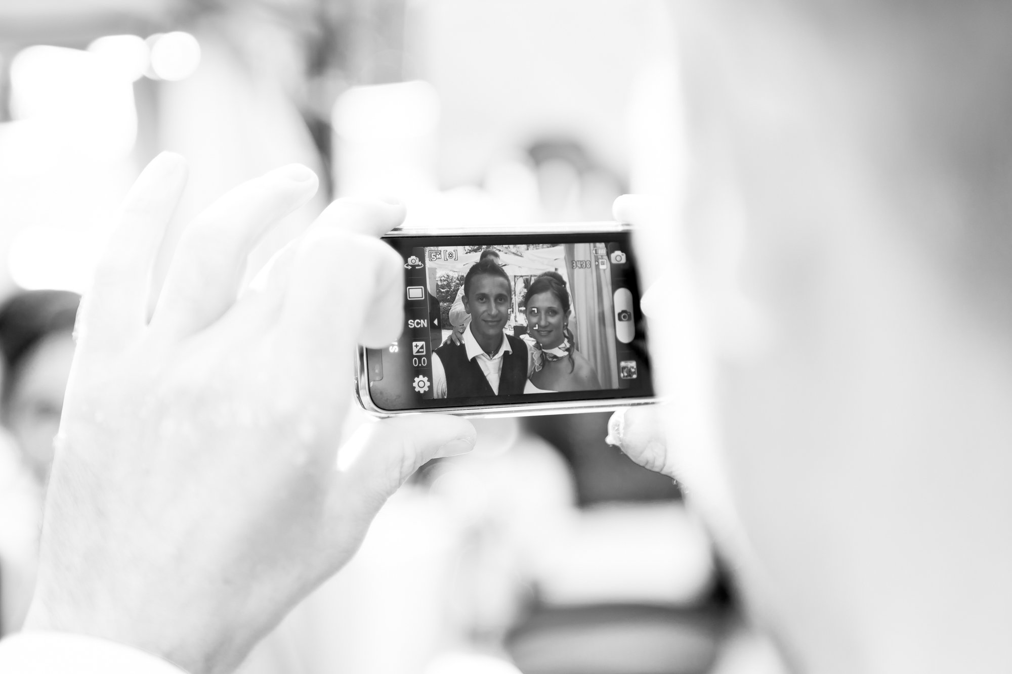 Person taking a black-and-white photo of a man and woman on a smartphone during a celebration or event.