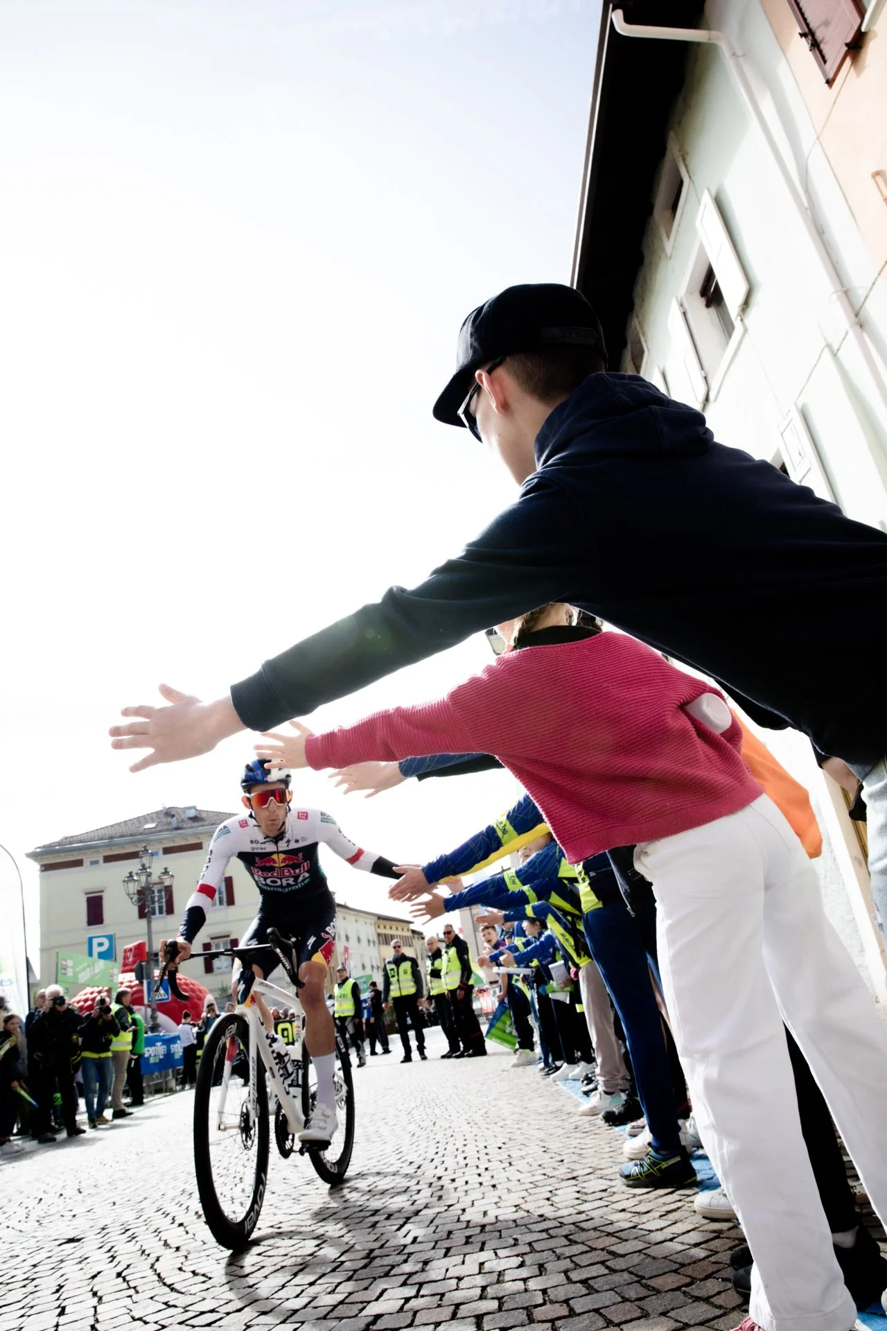 A cyclist in a White and red racing jersey and black shorts riding a mountain bike through a crowd who are reaching out to high five him, on a cobblestone street in an urban area.