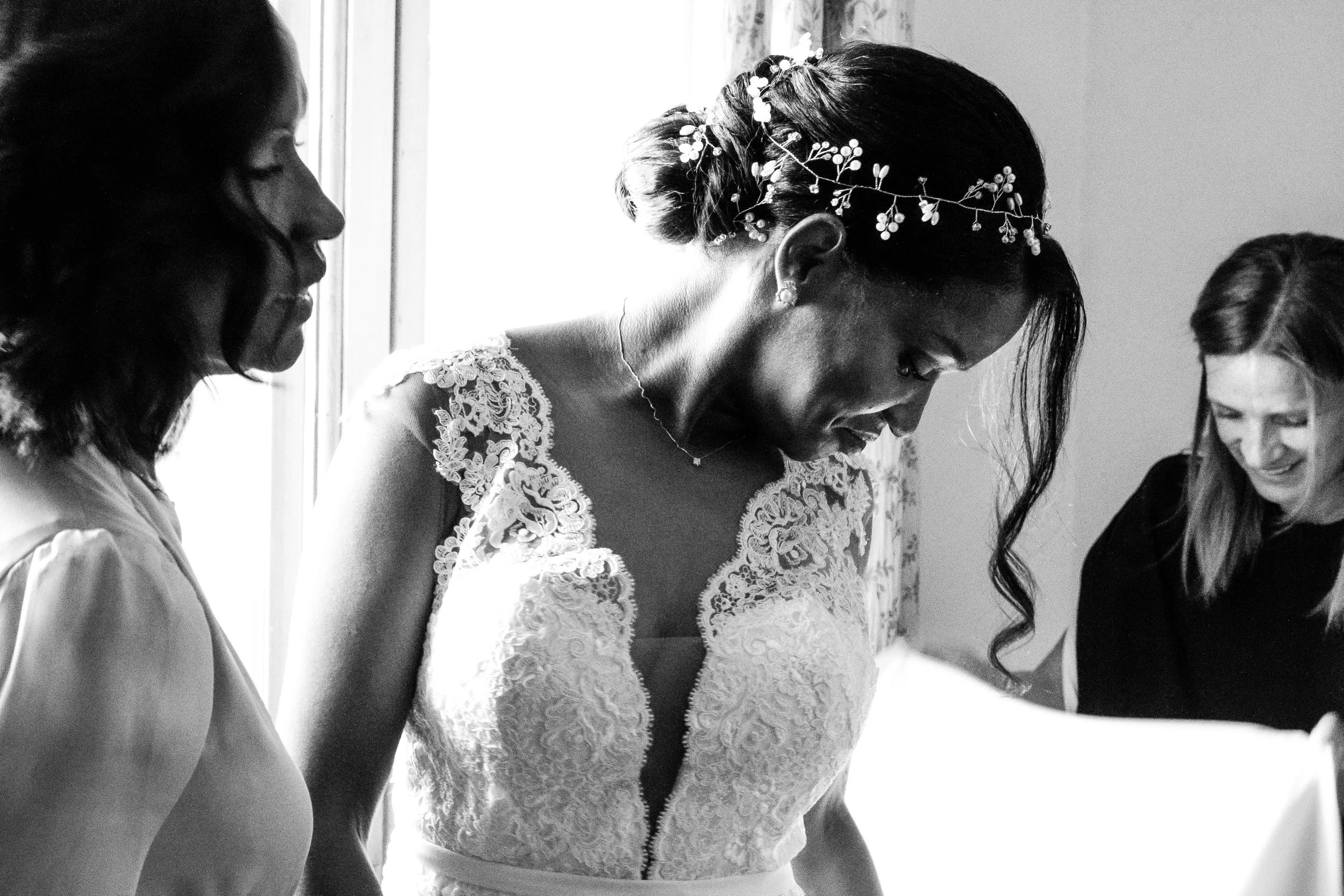 Black and white photo of three women, with one in a wedding dress, smiling while looking down, and two others standing beside her, in a room with a window.
