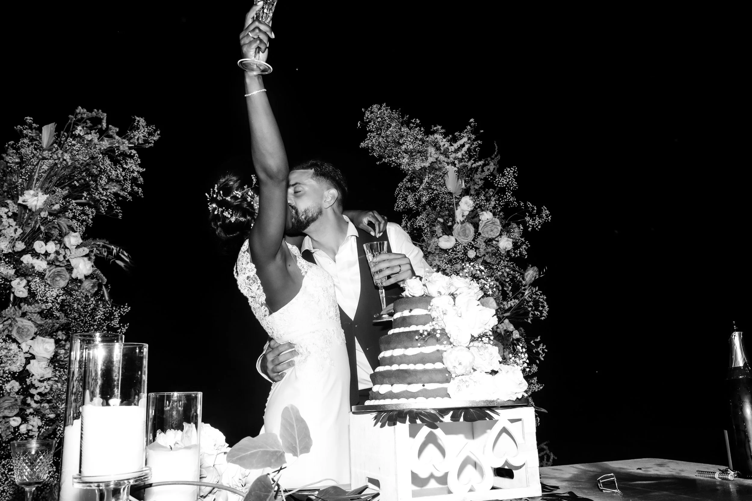 A black and white photo of a newlywed couple sharing a kiss during their wedding reception, the bride holding a glass of champagne with her arm raised, surrounded by wedding decorations, flowers, and a wedding cake.