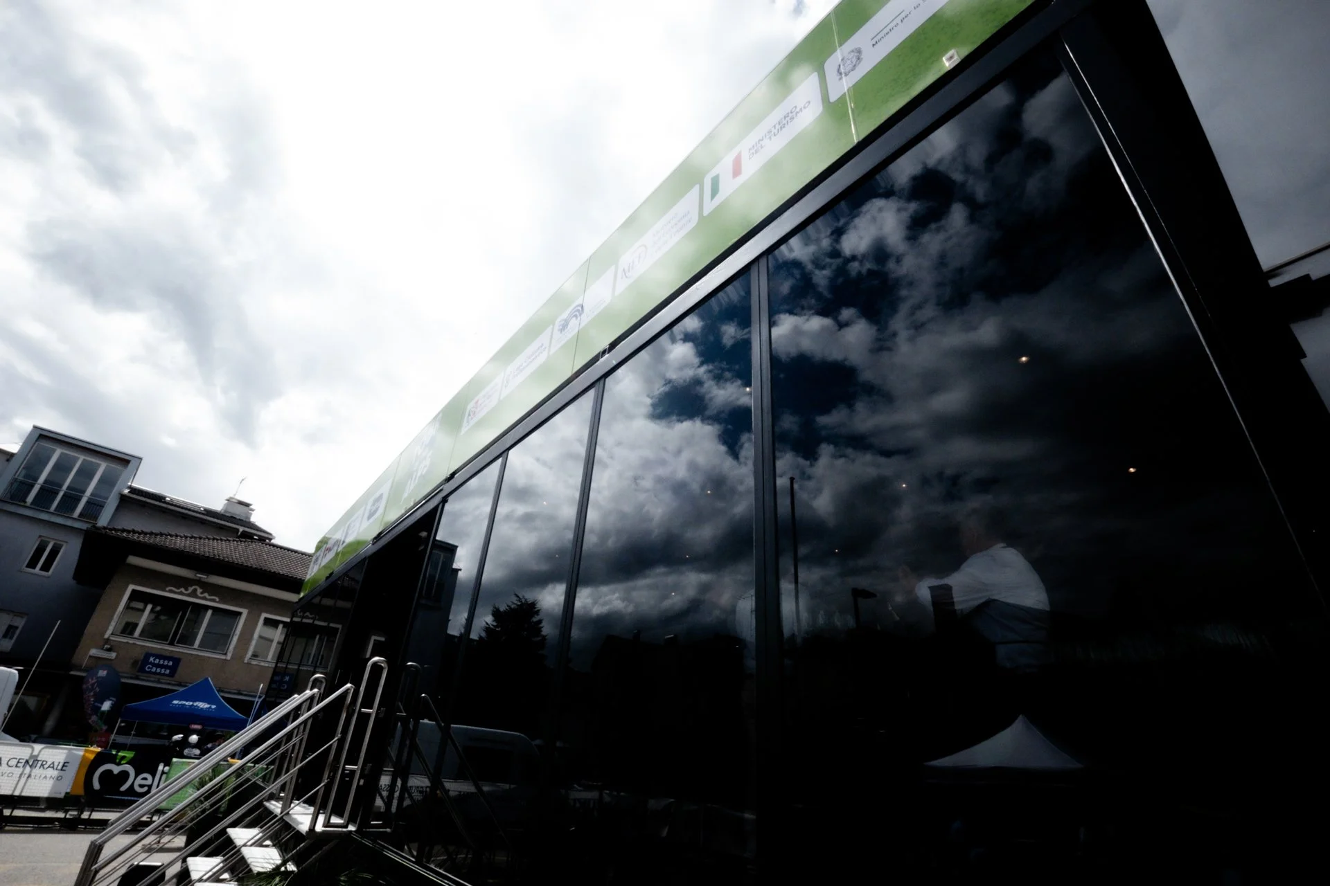 Reflection of cloudy sky on the glass facade of a modern building with street scene and tents in background.