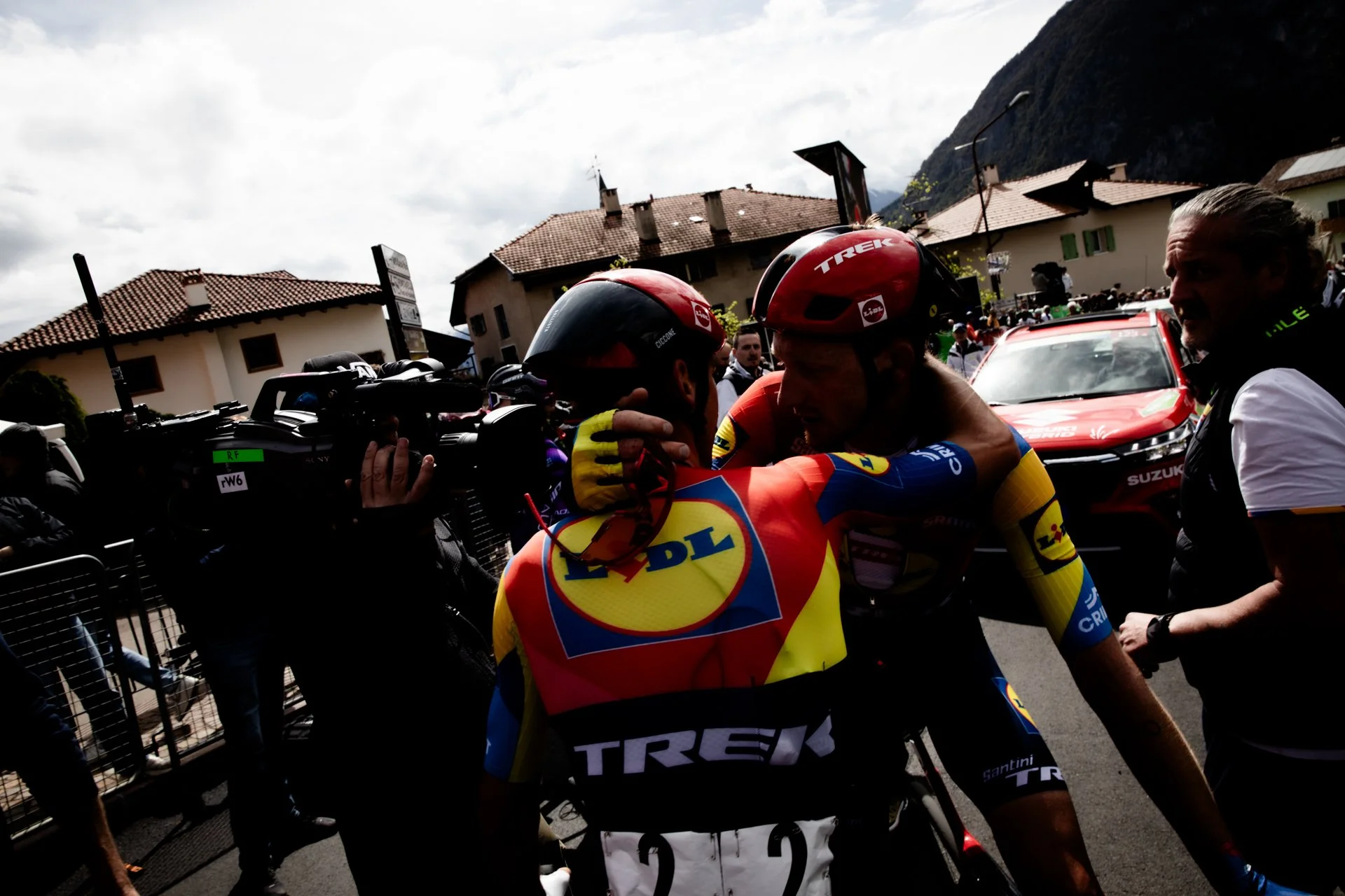 Two professional cyclists from Team Trek embracing after a race, surrounded by photographers and support staff, with parked cars and buildings in the background.