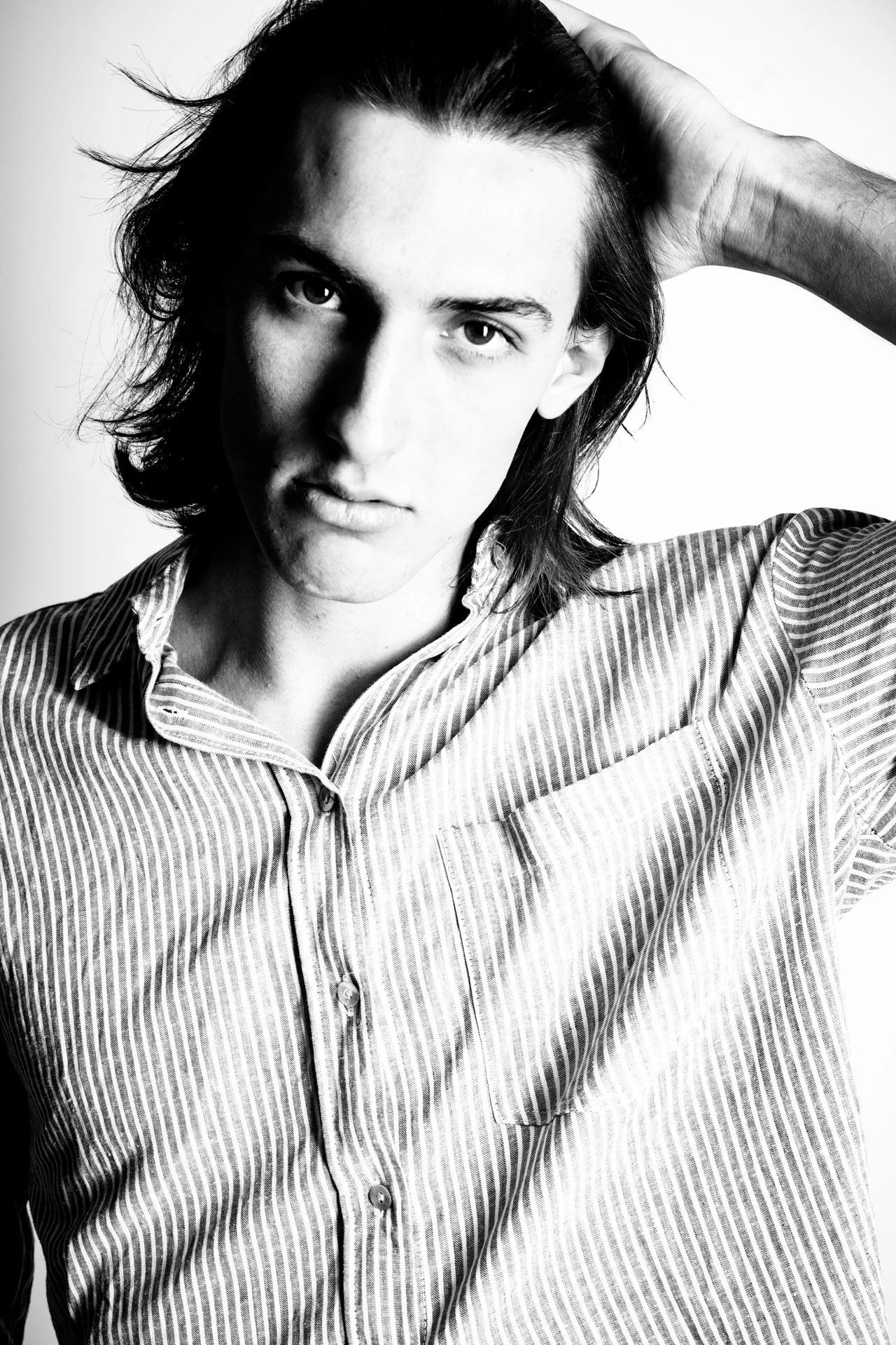 Black and white portrait of a young man with long hair, wearing a striped shirt, with one hand on his head and a serious expression.