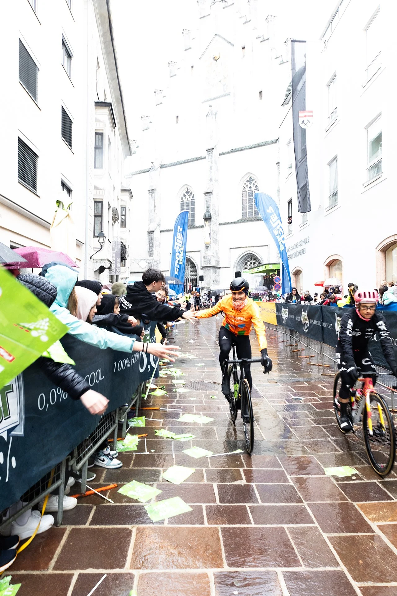 Cyclists participating in a race, with one cyclist in an orange shirt reaching out to high-five spectators along a wet street lined with barriers and spectators holding umbrellas, in front of a historic building with a church on a rainy day.