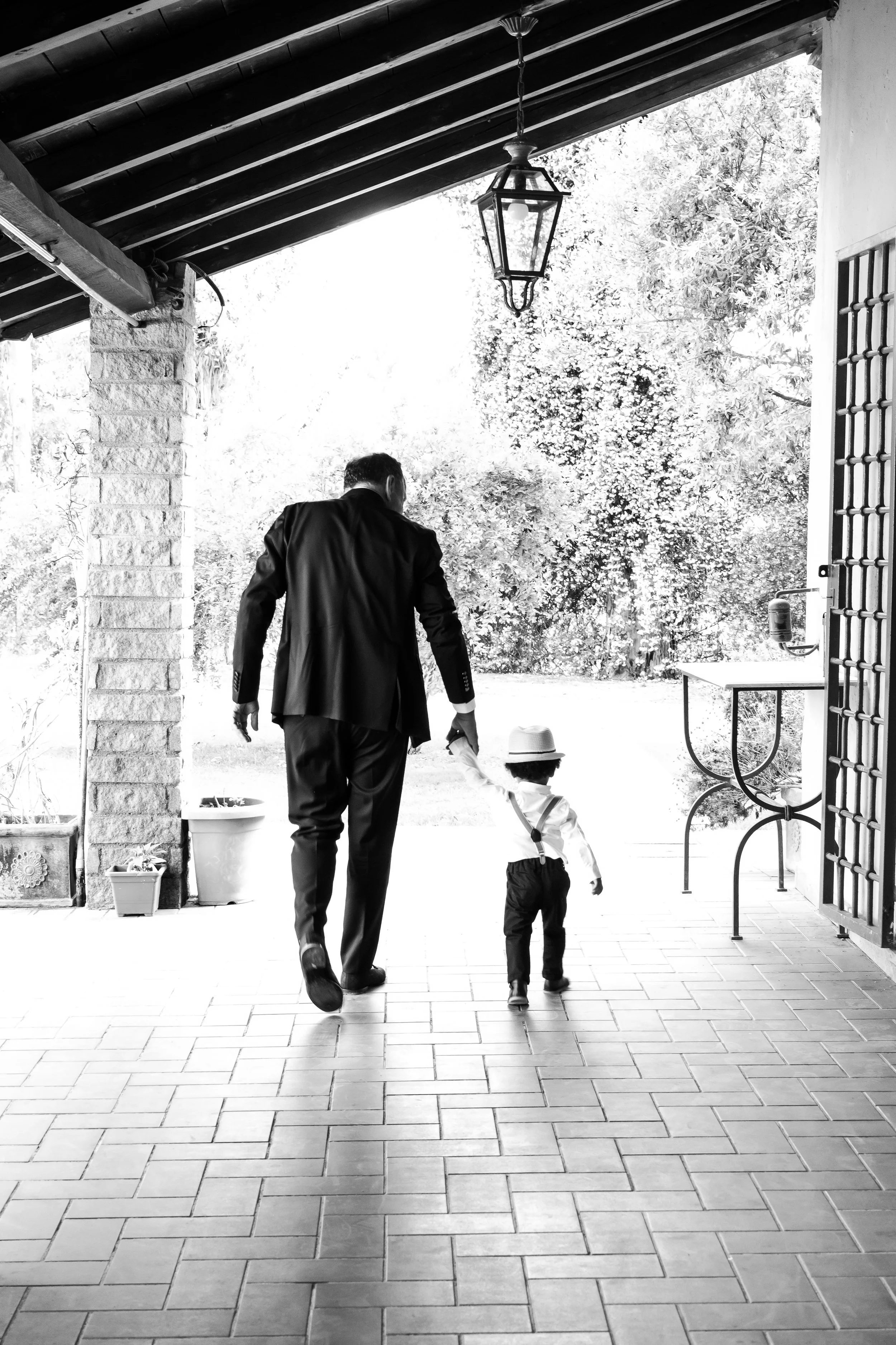 A man and a young boy, possibly father and son, walking hand in hand on a tiled patio. The man is dressed in a suit, and the boy is wearing a hat and suspenders. The patio is shaded by a roof with a hanging lantern, and there are trees and outdoor fu