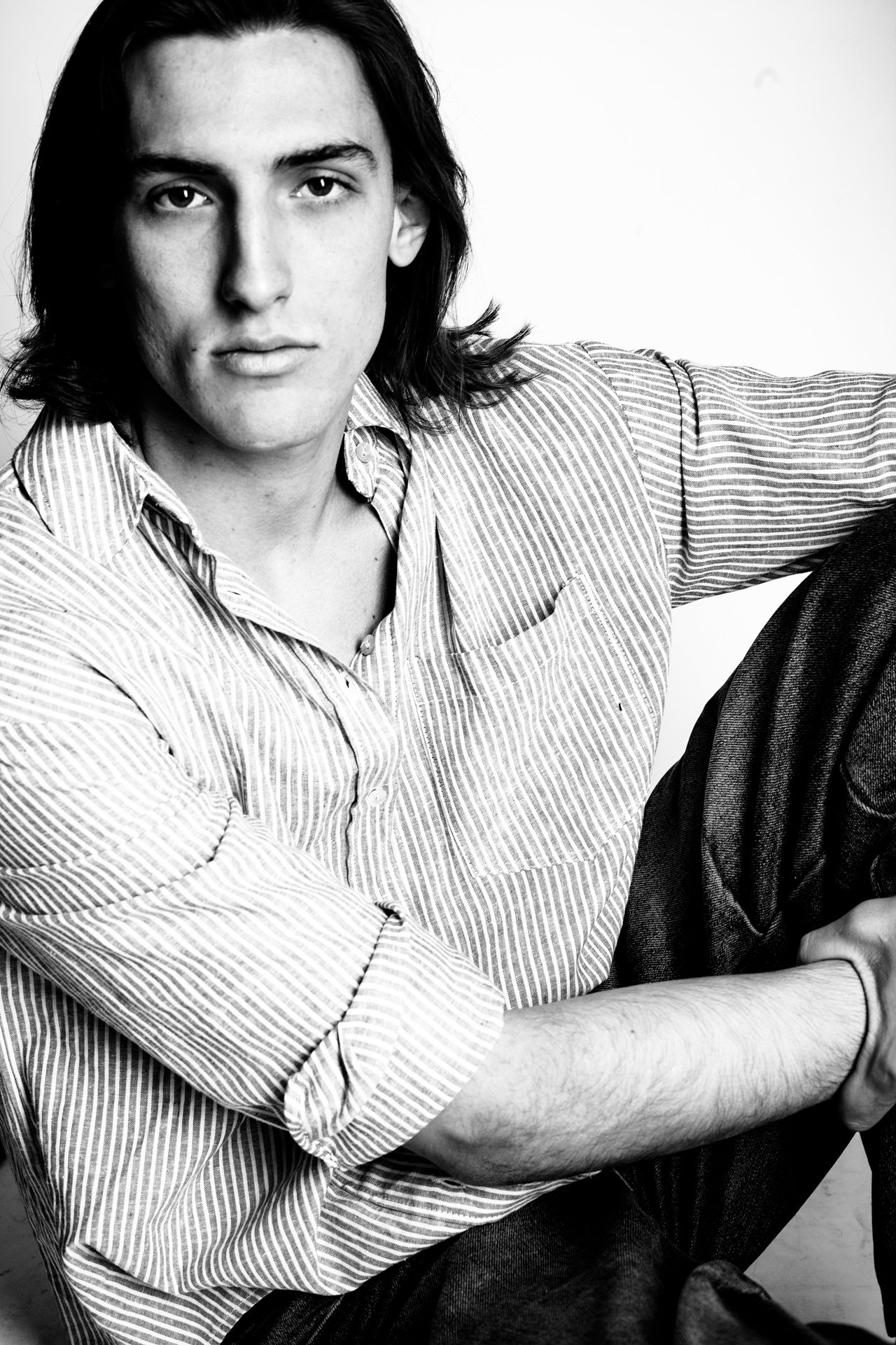 Black and white portrait of a young man with long, dark hair, wearing a striped button-up shirt, sitting with one knee up, looking directly at the camera with a serious expression.
