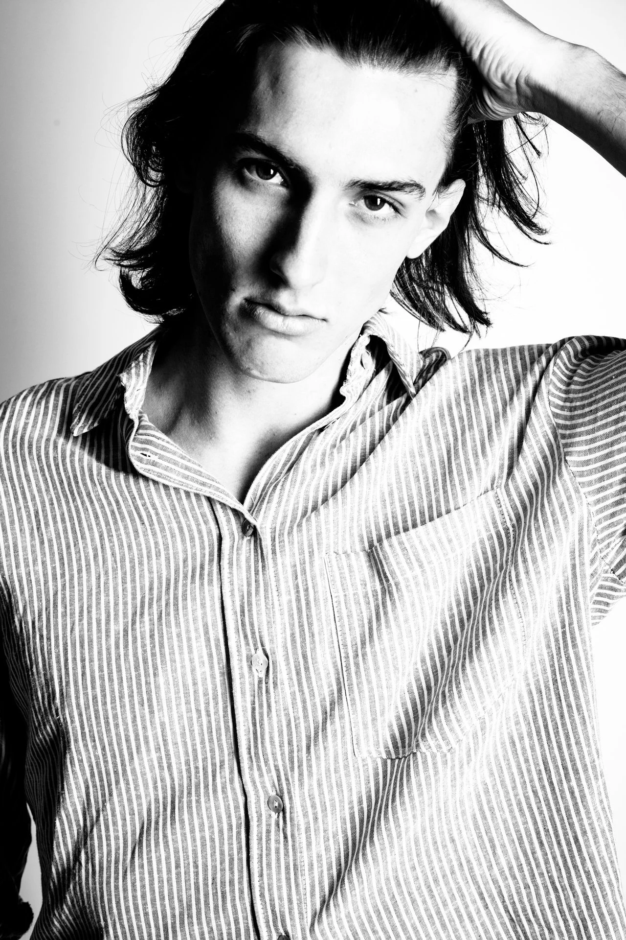 Black and white portrait of a young man with shoulder-length dark hair, wearing a striped button-up shirt, with one hand touching his hair and a serious expression.