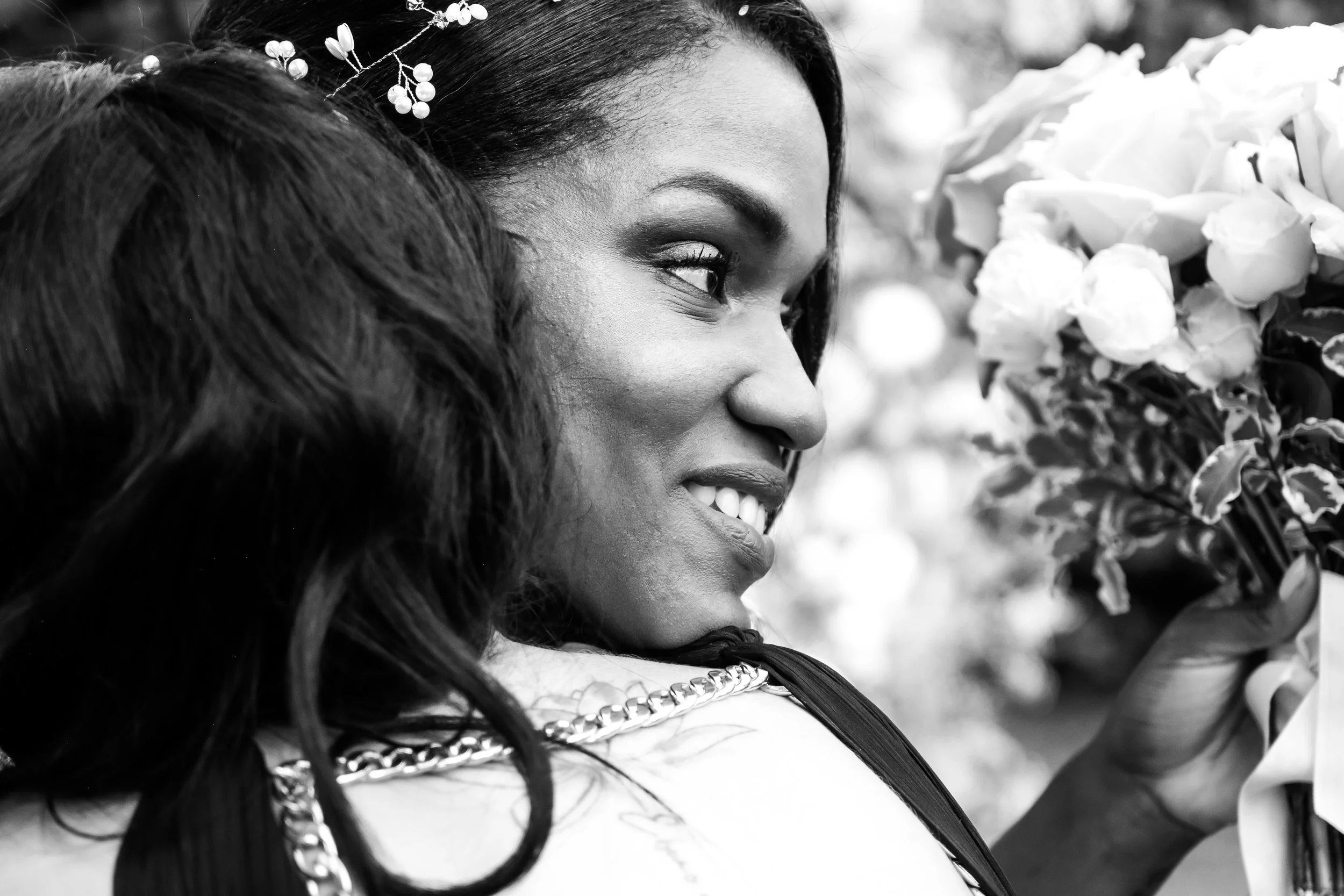 A woman with long dark hair and a decorative hairpiece holds a large bouquet of roses, smiling softly. The photo is in black and white.