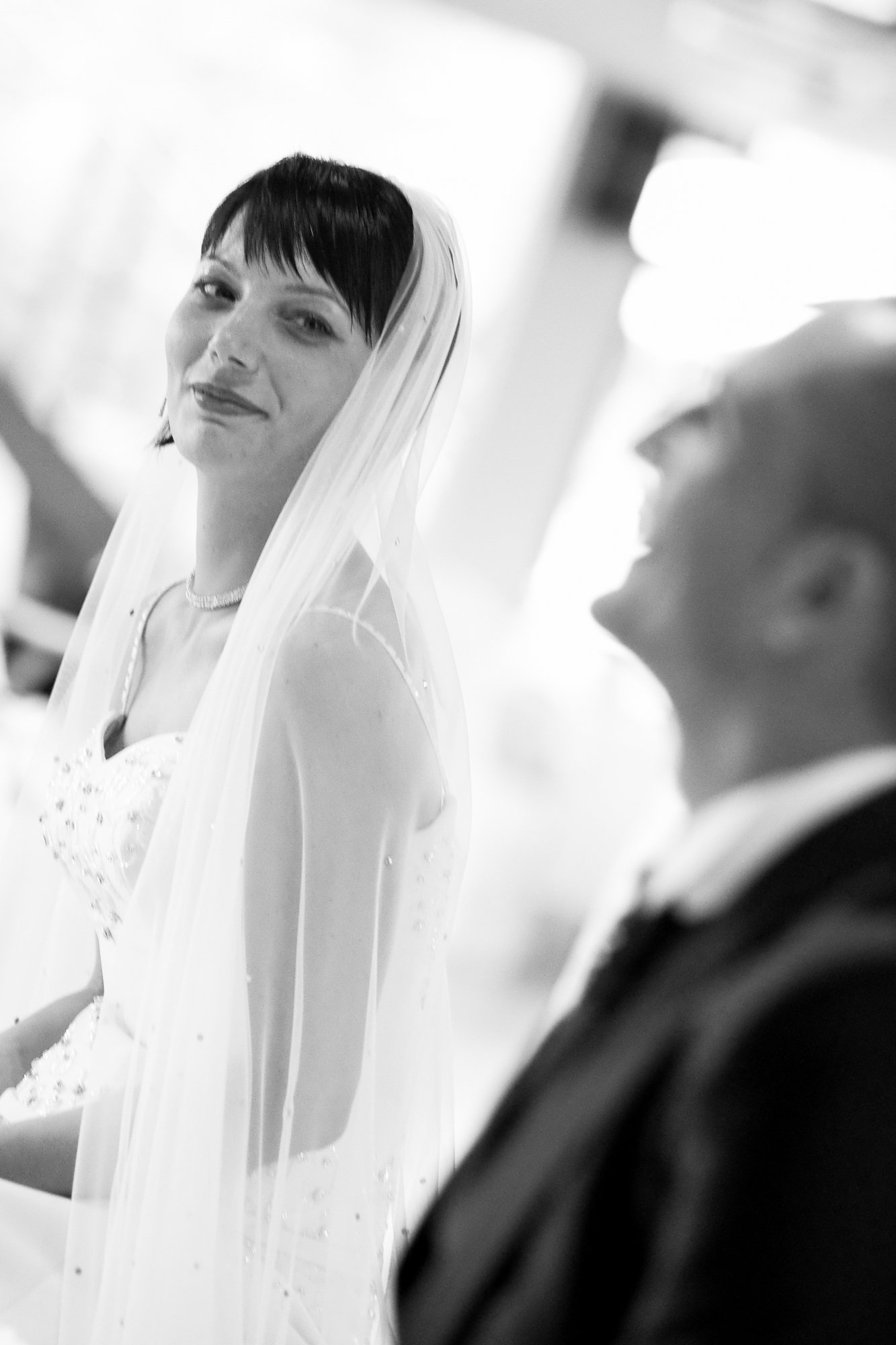 A black-and-white photo of a woman in a wedding dress with a veil, smiling gently, and a blurred man in a suit beside her, at a wedding ceremony.