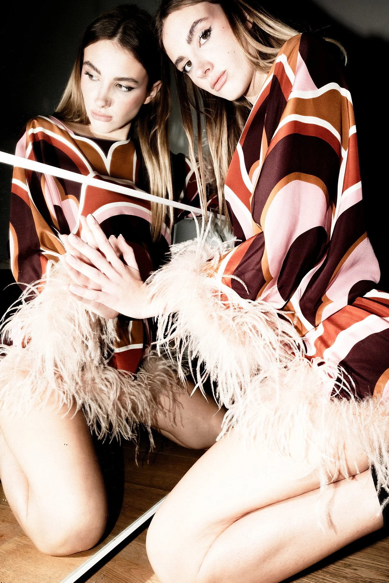 Two women in patterned dresses kneel on the floor, looking into a mirror, with one woman touching her hand on the other woman's arm, and feathered cuffs on their sleeves.