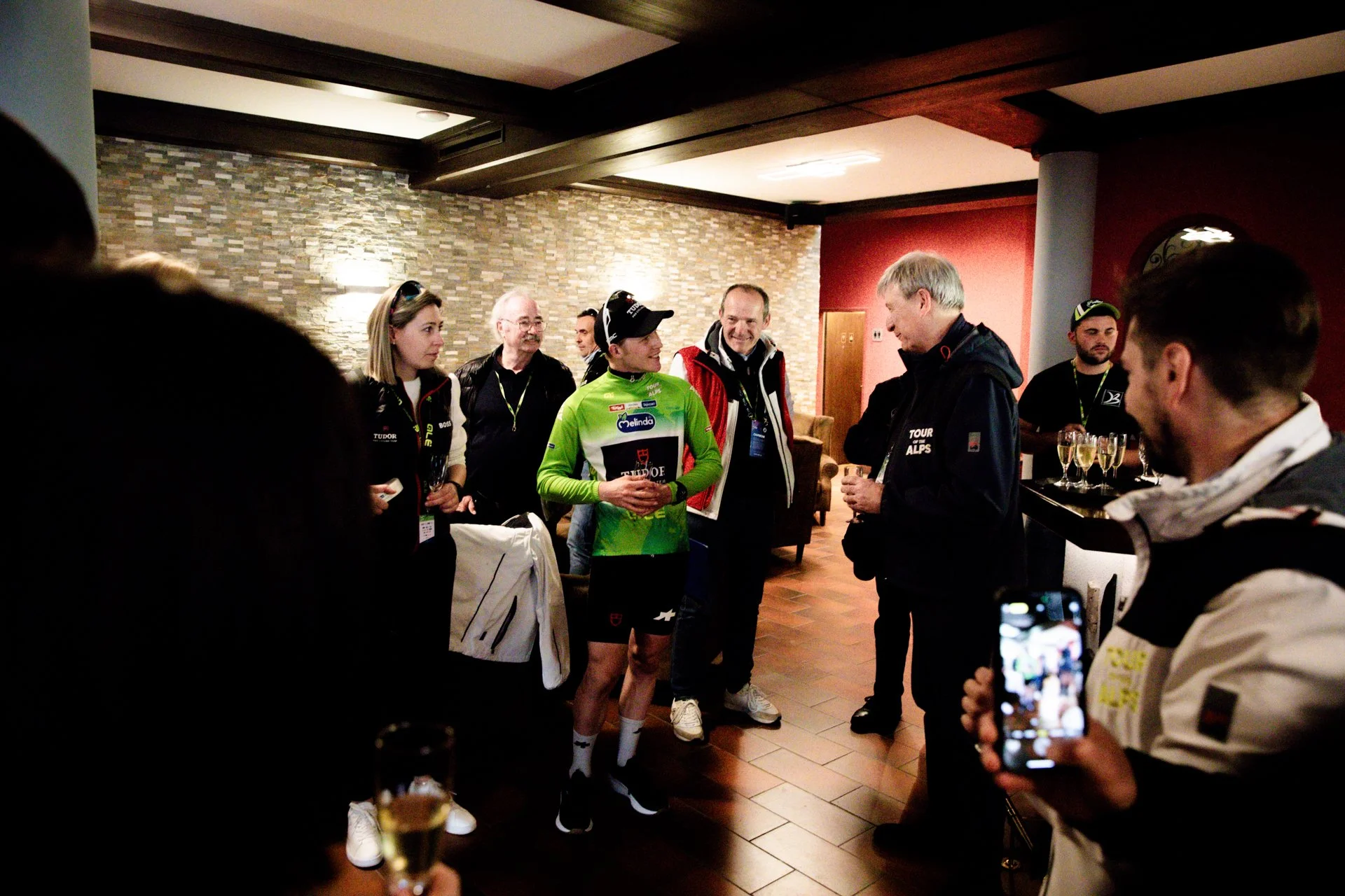 Group of people socializing at an indoor event, some holding glasses of champagne, with a brick wall and red wall in the background.